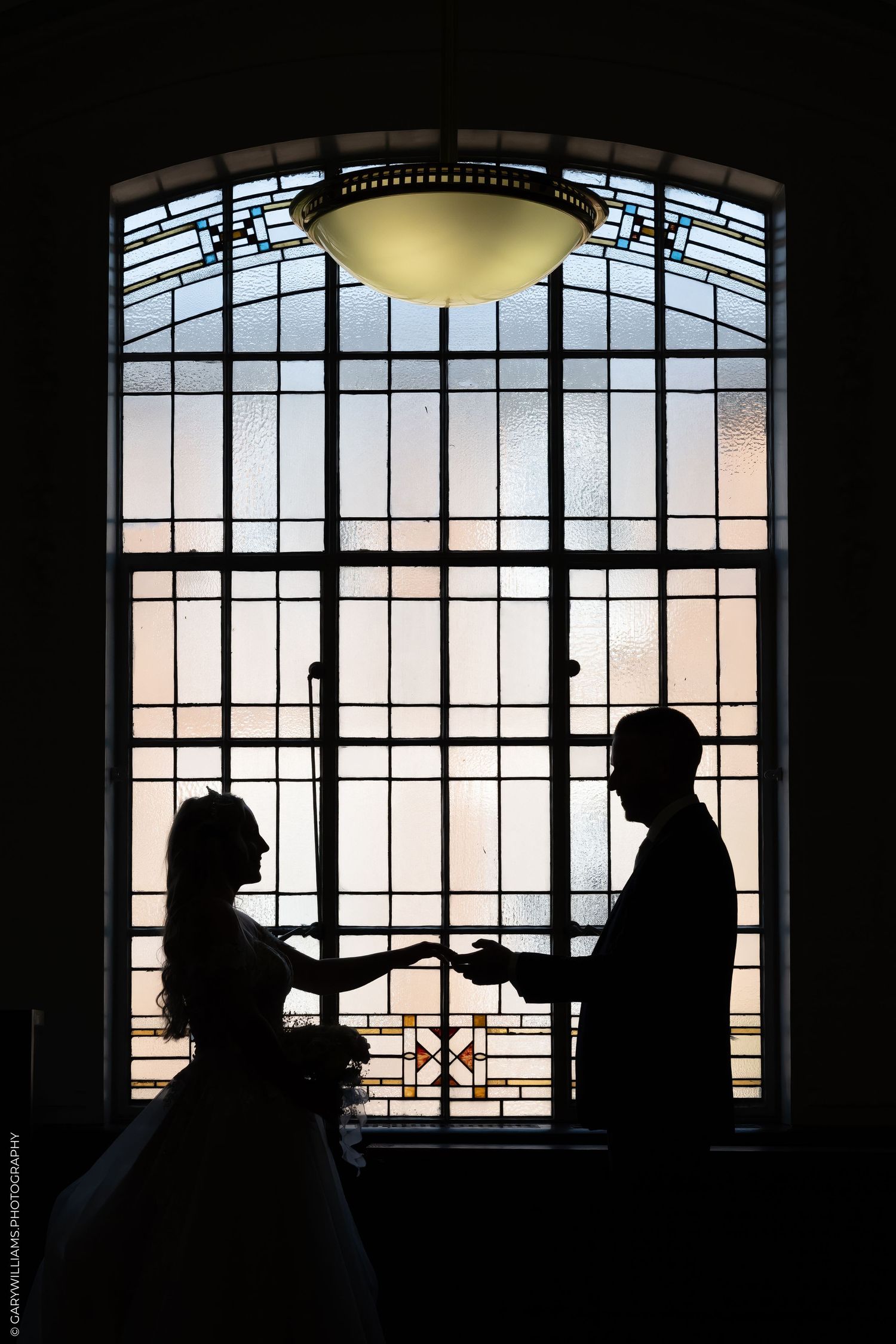 Silhouette of bride and groom holding hands before a stunning stained glass arched window at golden hour.