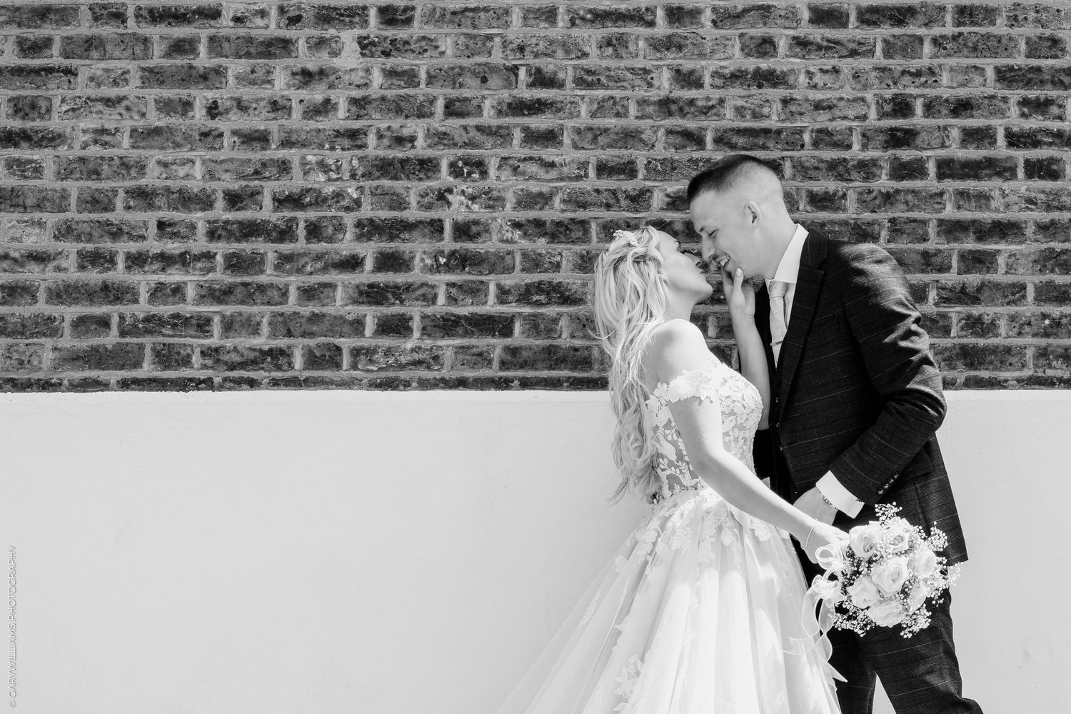 Black and white photo of a bride and groom holding hands and leaning in close against a brick wall.