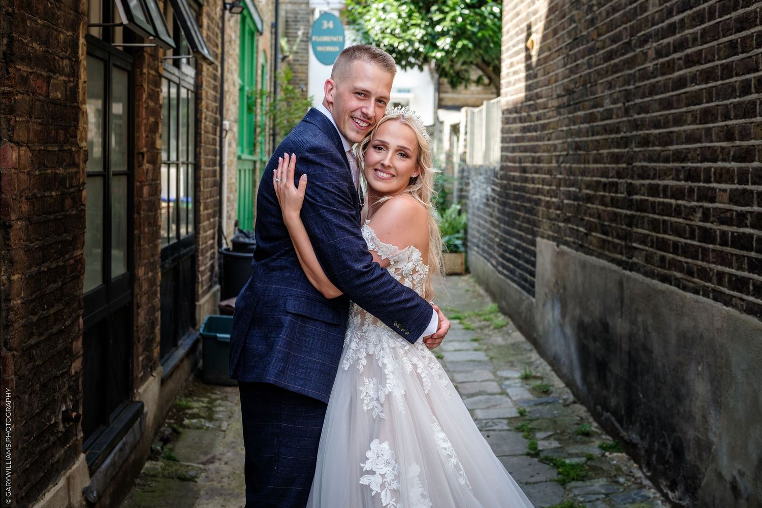 A bride and groom embrace in a charming brick alleyway, smiling joyfully on their wedding day.