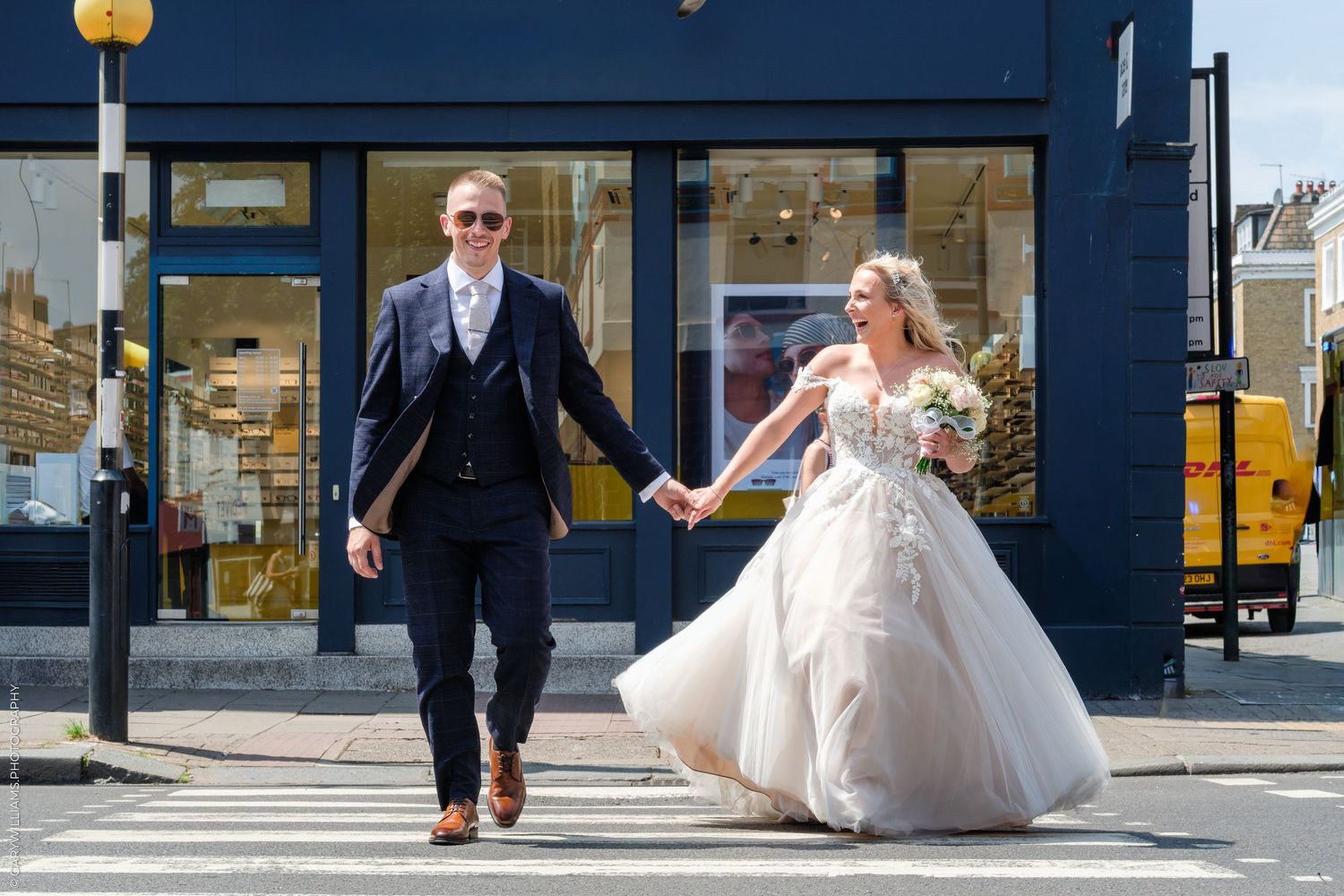 Newlywed couple holding hands crossing street, bride in ballgown, groom in navy suit, on sunny day.