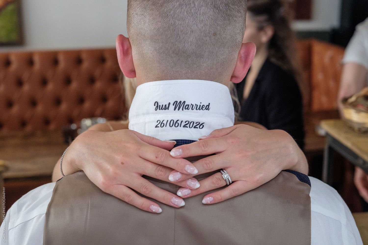 Bride hugging groom from behind, showing 'Just Married 26/06/2026' embroidered on his white collar.