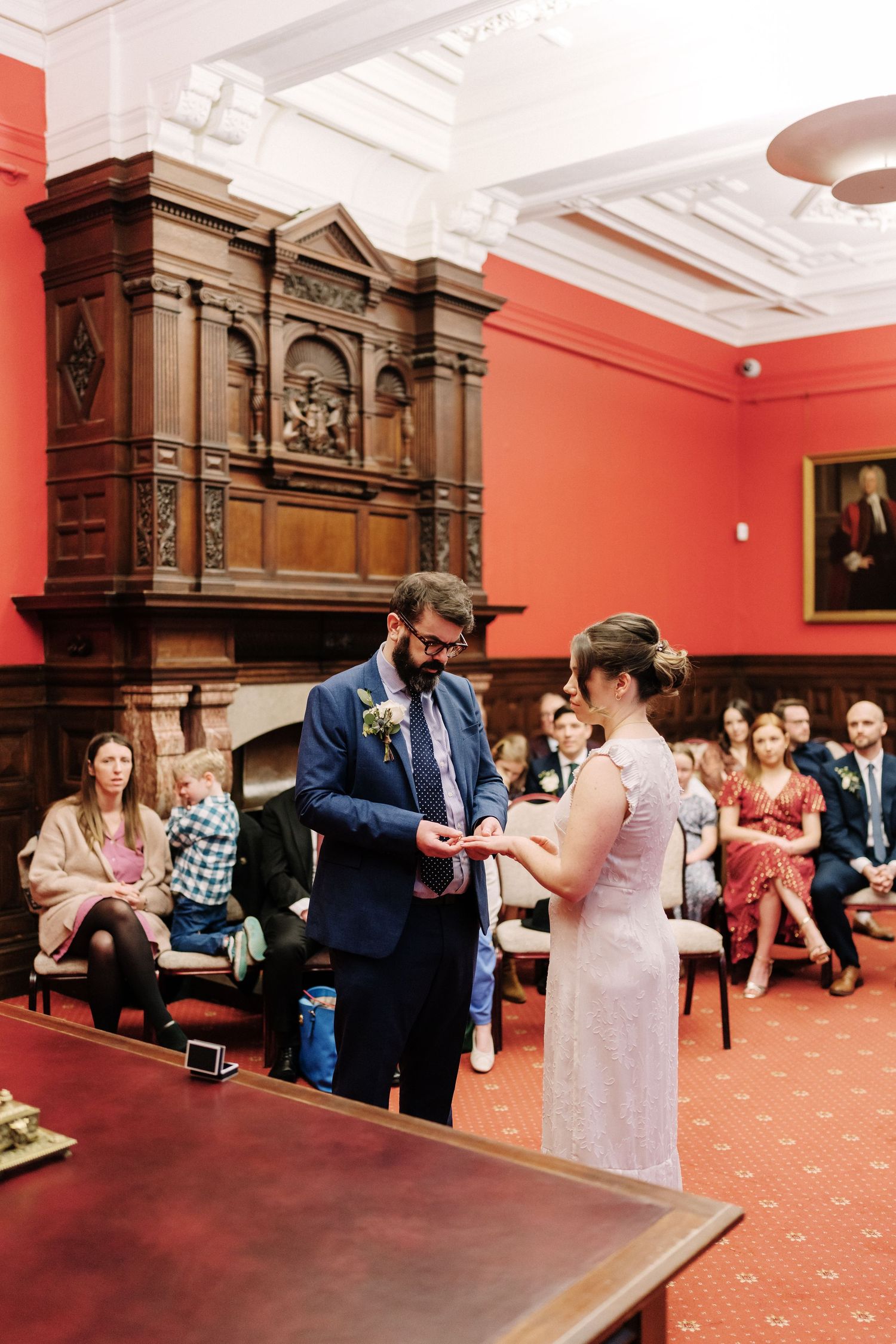 Couple exchanging rings during wedding ceremony in elegant red-walled venue with ornate wooden fireplace and seated guests.