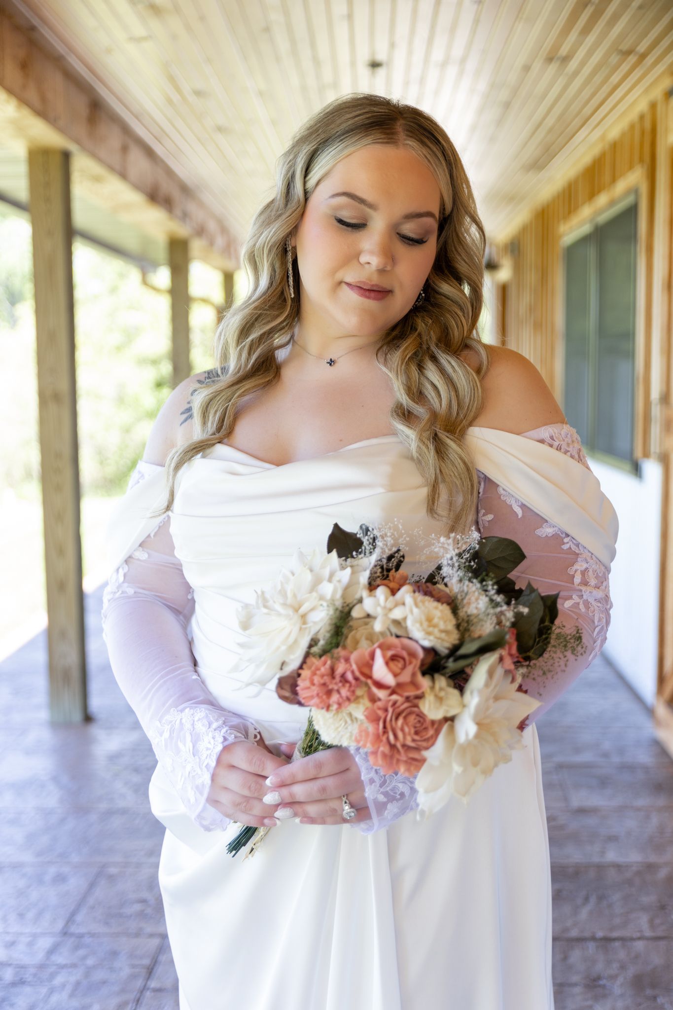 Bride in off-shoulder white gown with lace sleeves holds a bouquet of pink and cream roses outdoors.