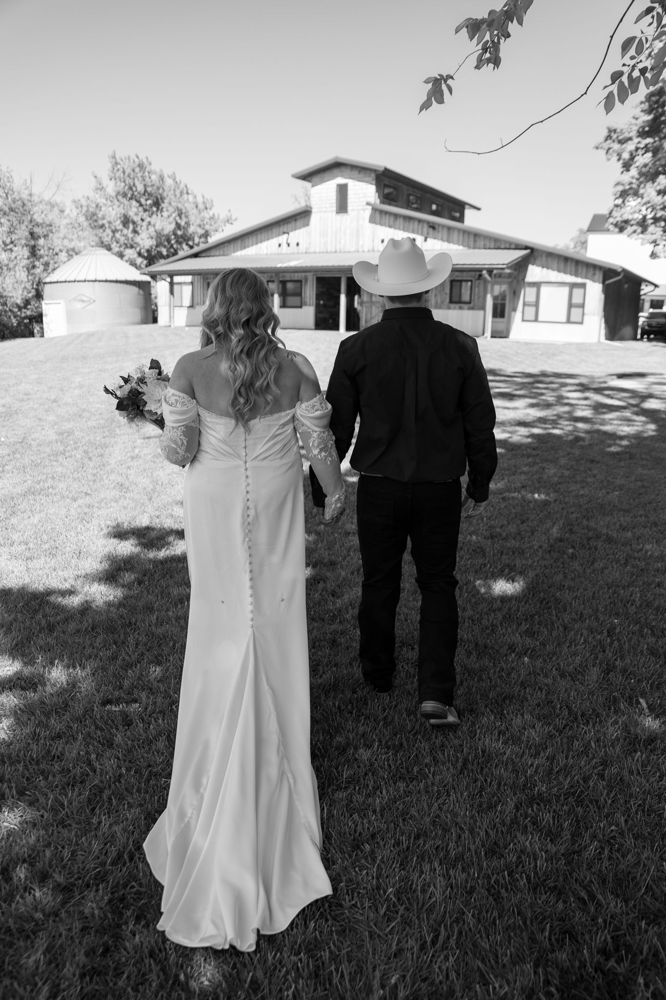 Black and white photo of bride and groom holding hands walking toward a rustic barn venue, groom wearing cowboy hat.