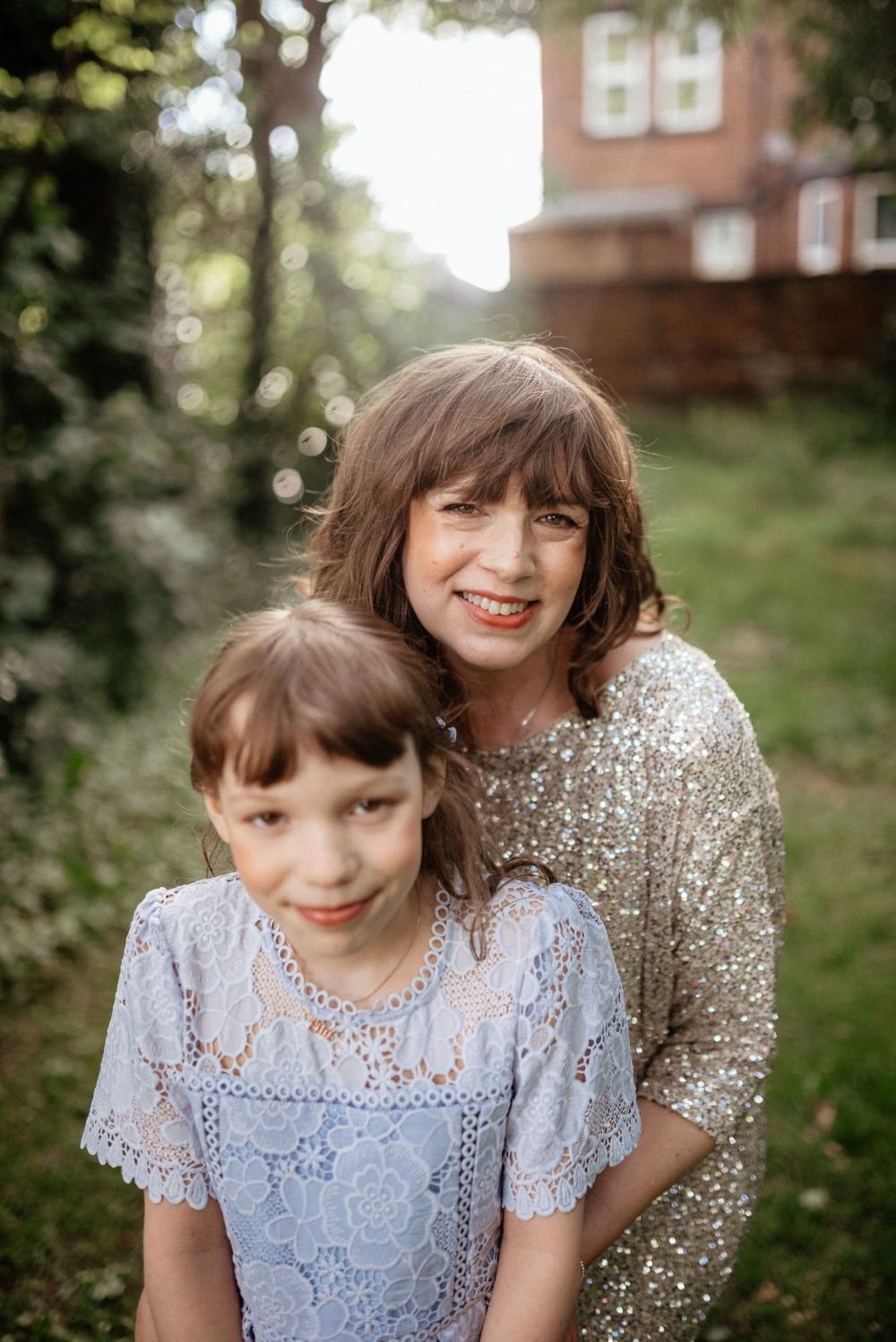 Mother and daughter in sparkly and lace dresses share a joyful moment in a garden setting.