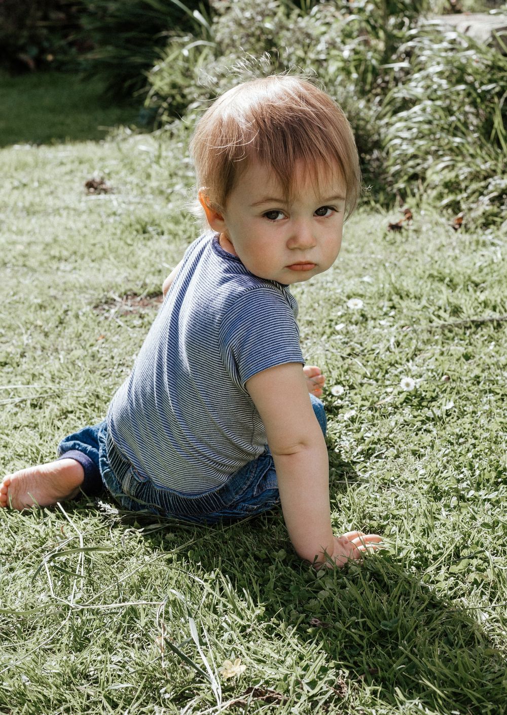 Baby in grey t-shirt and overalls crawling on sunny grass.
