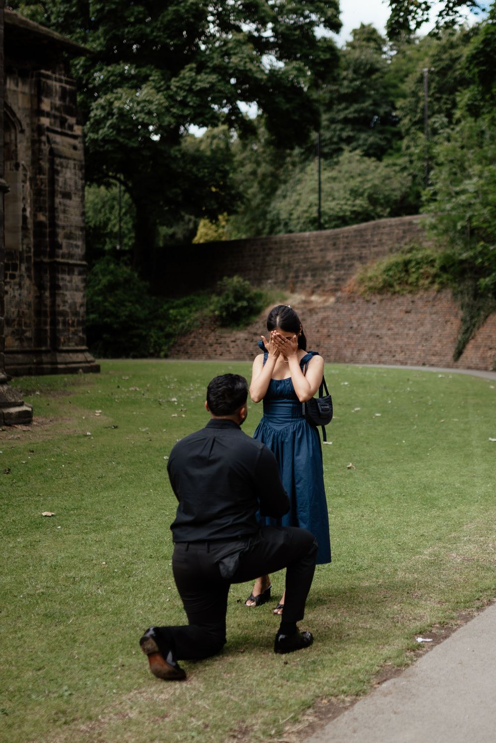 A romantic proposal scene on a grassy lawn next to stone walls.