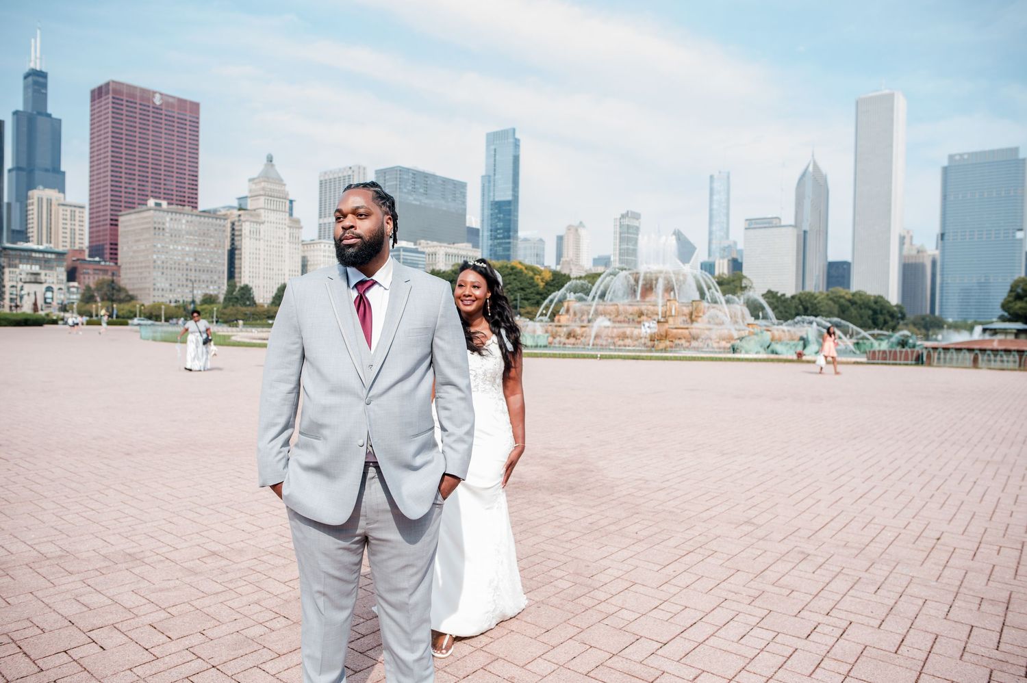 Couple in wedding attire poses near Buckingham Fountain with Chicago skyline backdrop on a sunny day.