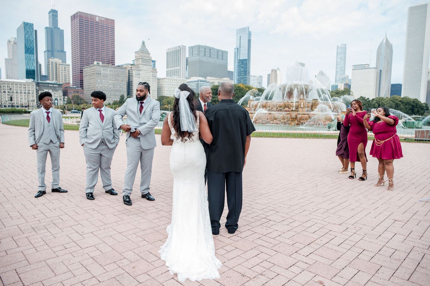 Outdoor wedding ceremony at Buckingham Fountain in Chicago with bridal party in gray suits and burgundy dresses.