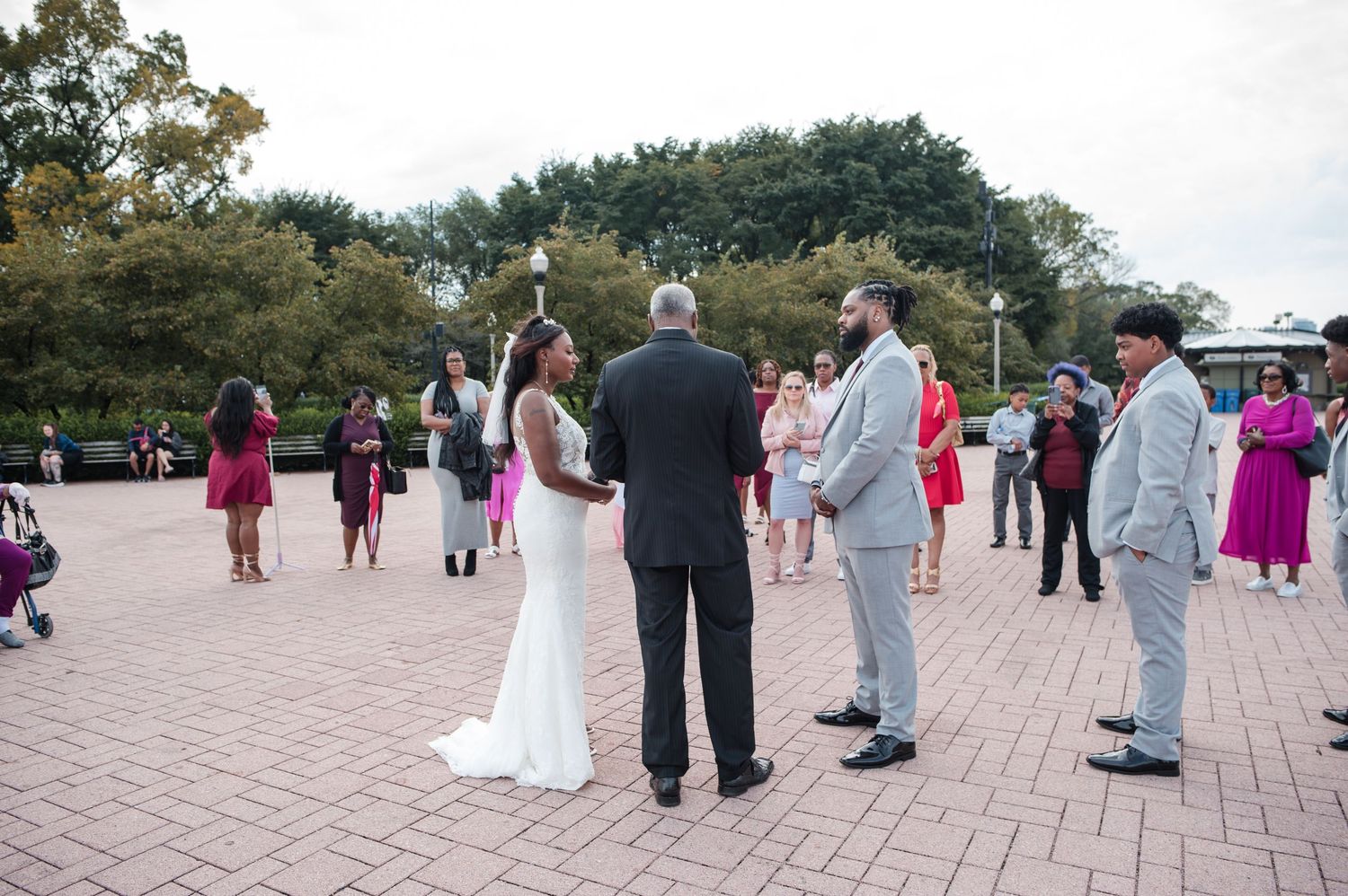Outdoor wedding ceremony on brick plaza with bride in white gown, groom, and officiant surrounded by guests.