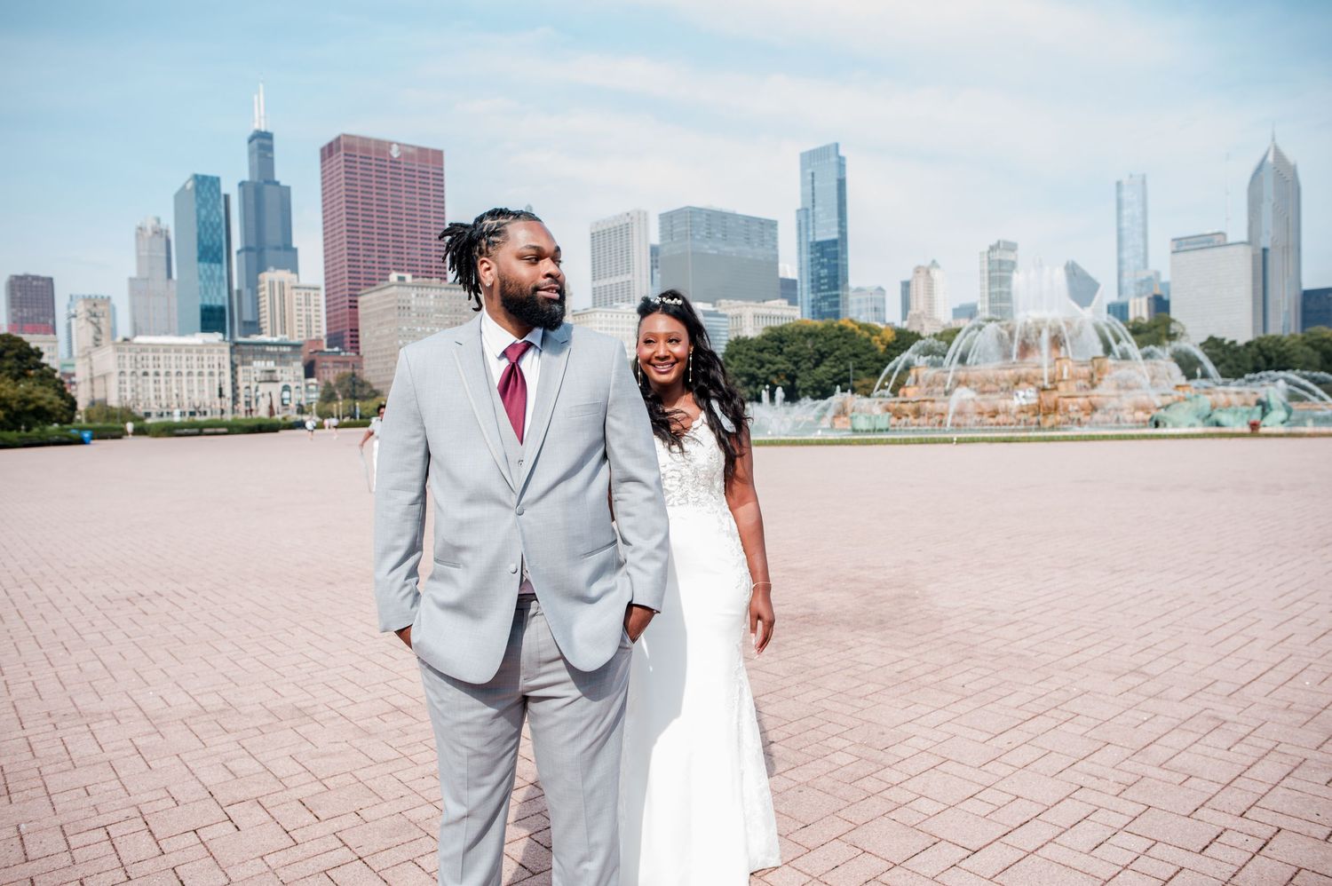 Couple in wedding attire poses near Buckingham Fountain with Chicago skyline backdrop on a sunny day.