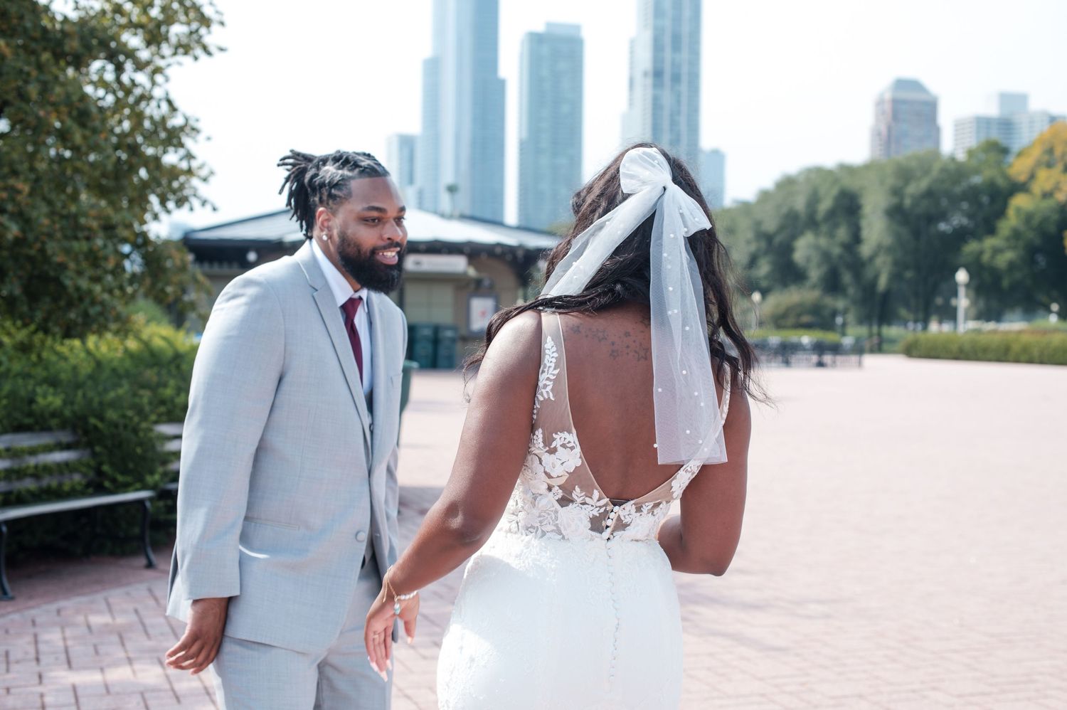 A bride and groom hold hands outdoors with a city skyline backdrop, the bride in a lace backless gown and veil.