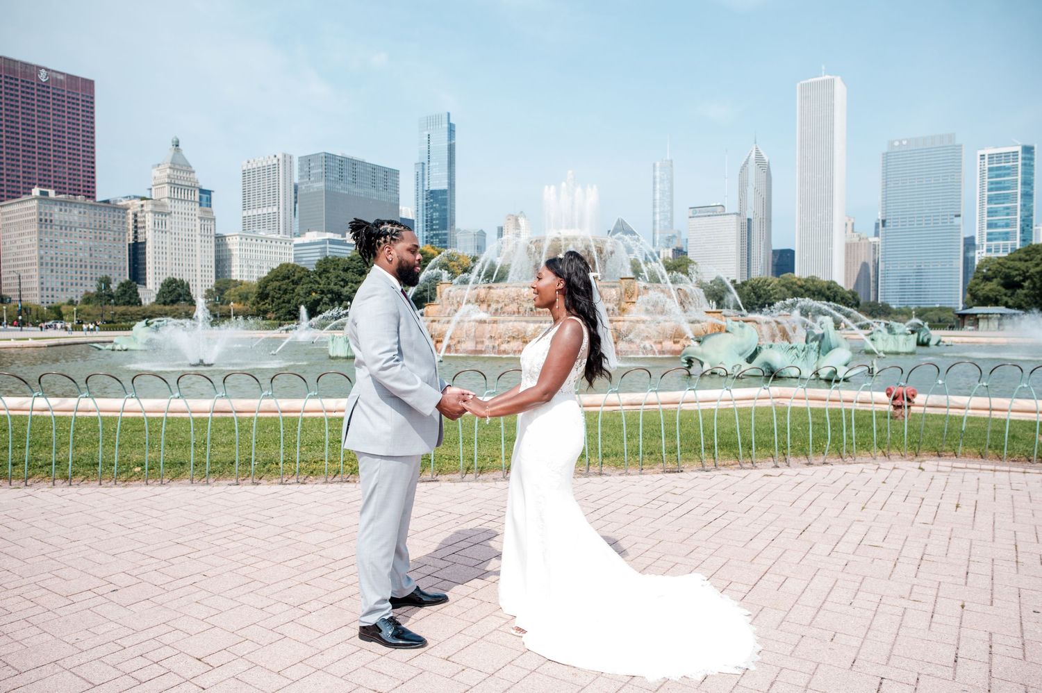 Newlyweds hold hands facing each other near Buckingham Fountain with Chicago city skyline behind them.