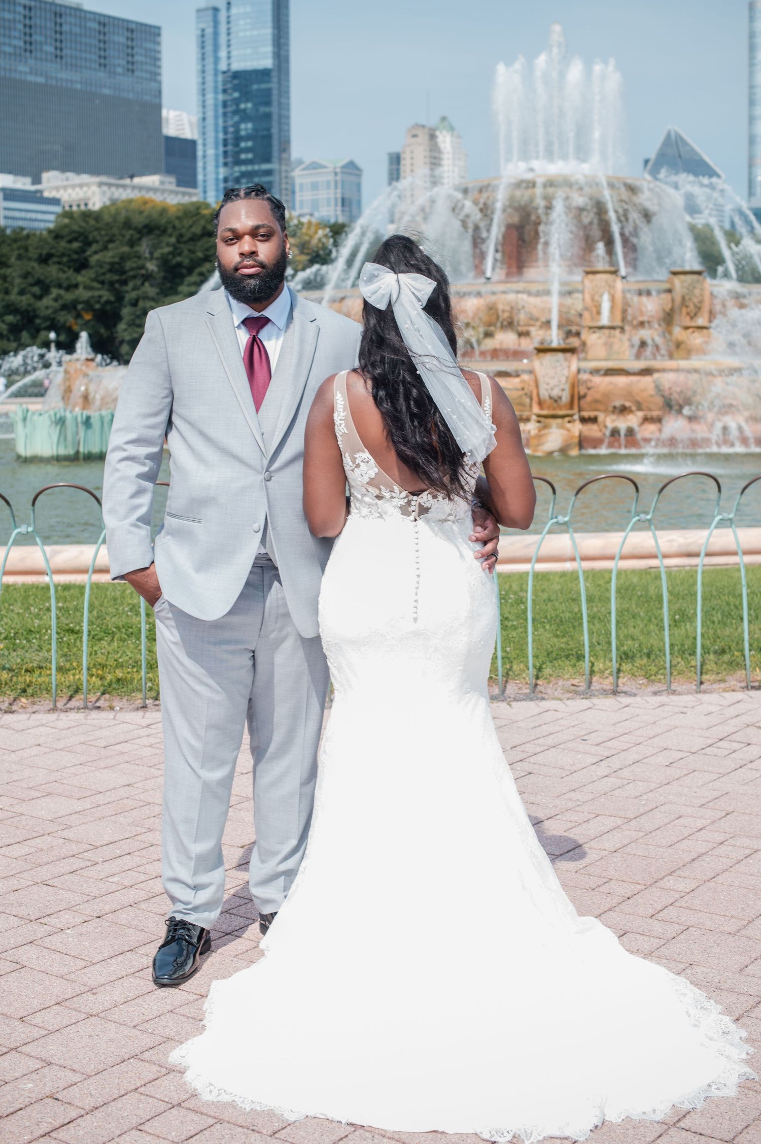 A bride and groom pose in front of Buckingham Fountain in Chicago, she in a white mermaid gown, he in a gray suit.