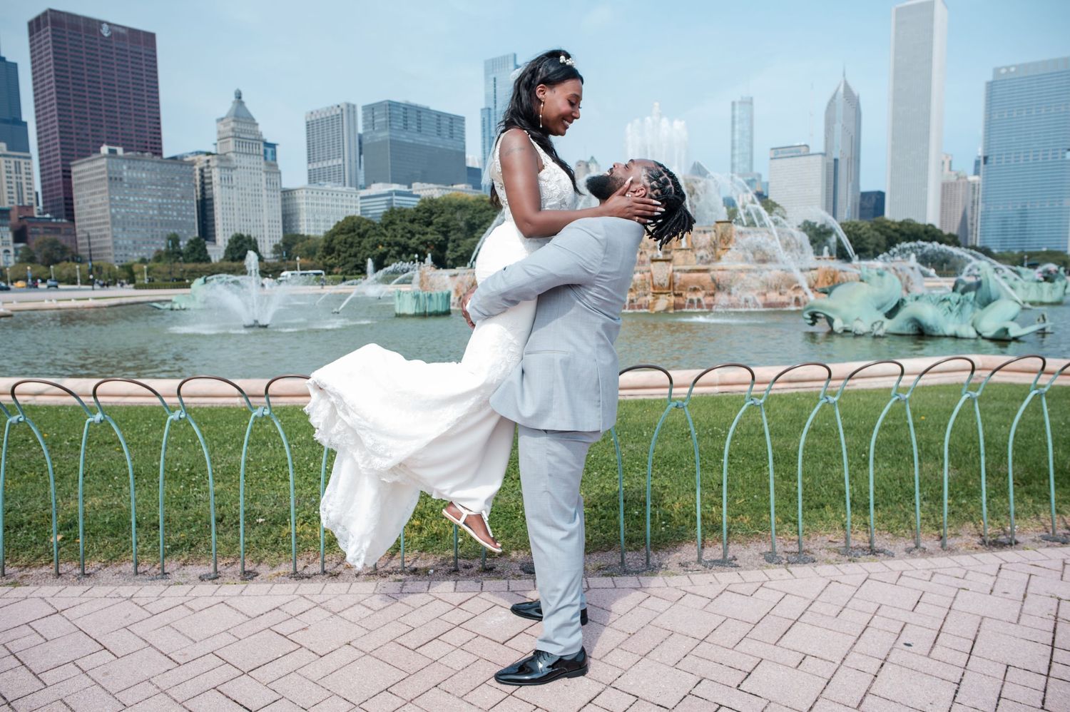 Groom lifts bride in white dress near Buckingham Fountain with Chicago skyline backdrop on their wedding day.