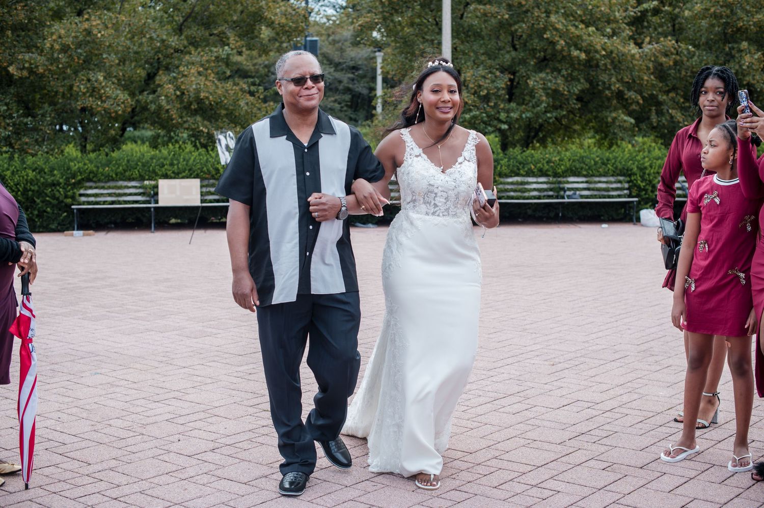A bride in a white gown walks with an older man in sunglasses at an outdoor wedding ceremony.