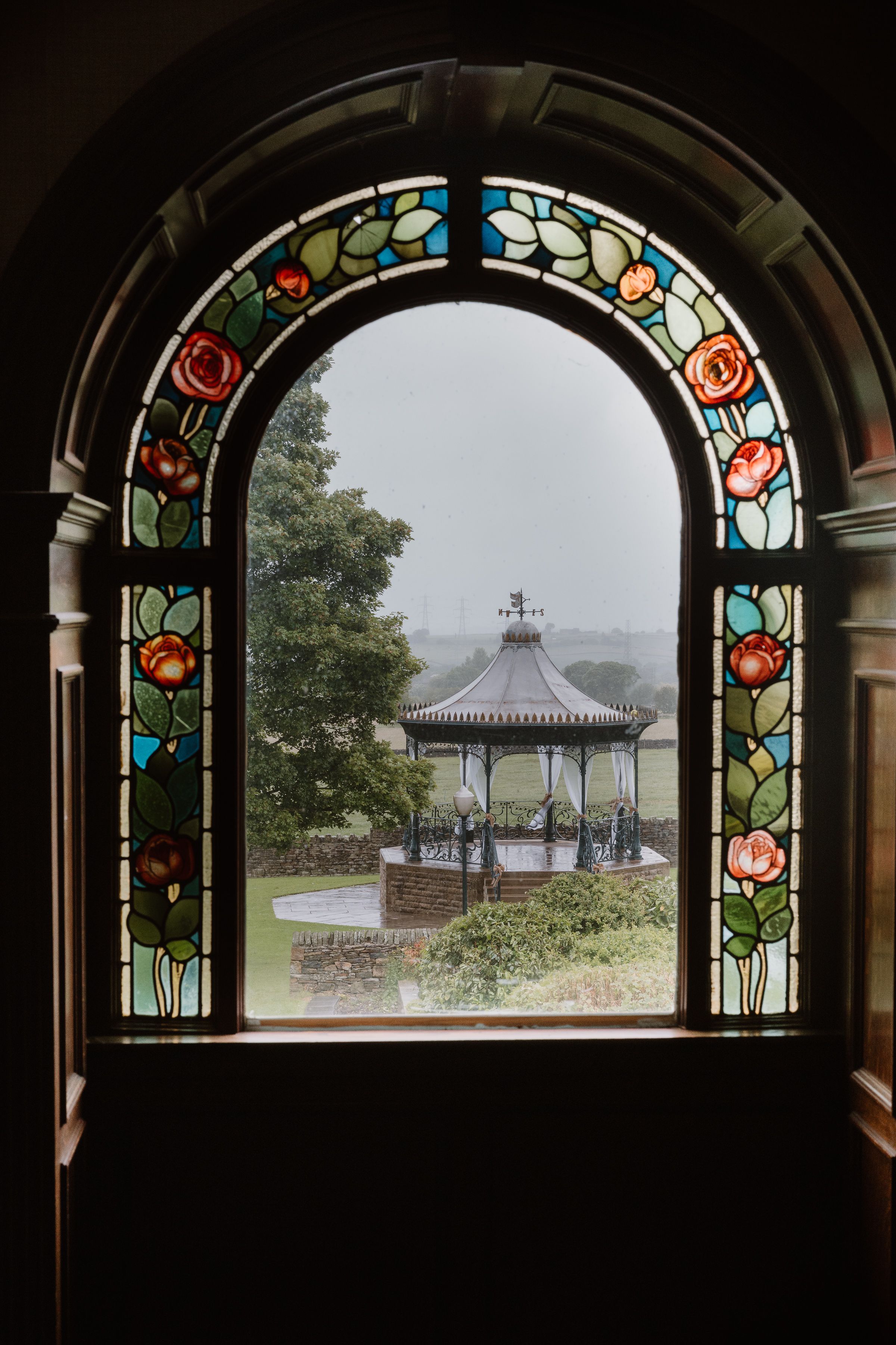 Stained glass arched window with roses framing a view of a Victorian bandstand in a misty park.