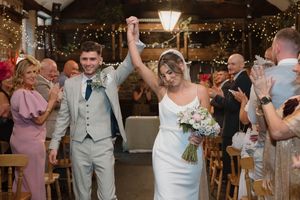 Joyful newlyweds raise hands walking down aisle at rustic barn wedding venue with fairy lights.
