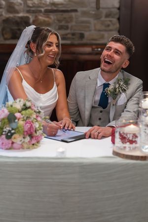Laughing bride and groom signing wedding register at decorated table with flowers and candles in stone venue.