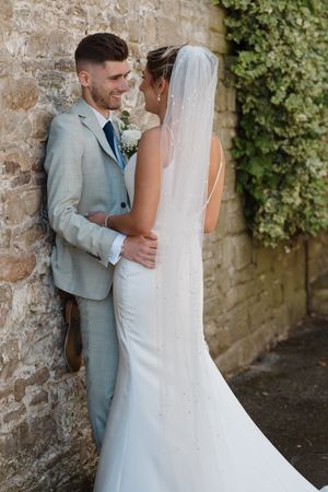 A smiling groom in a light gray suit embraces his bride in a white gown and veil against a stone wall.