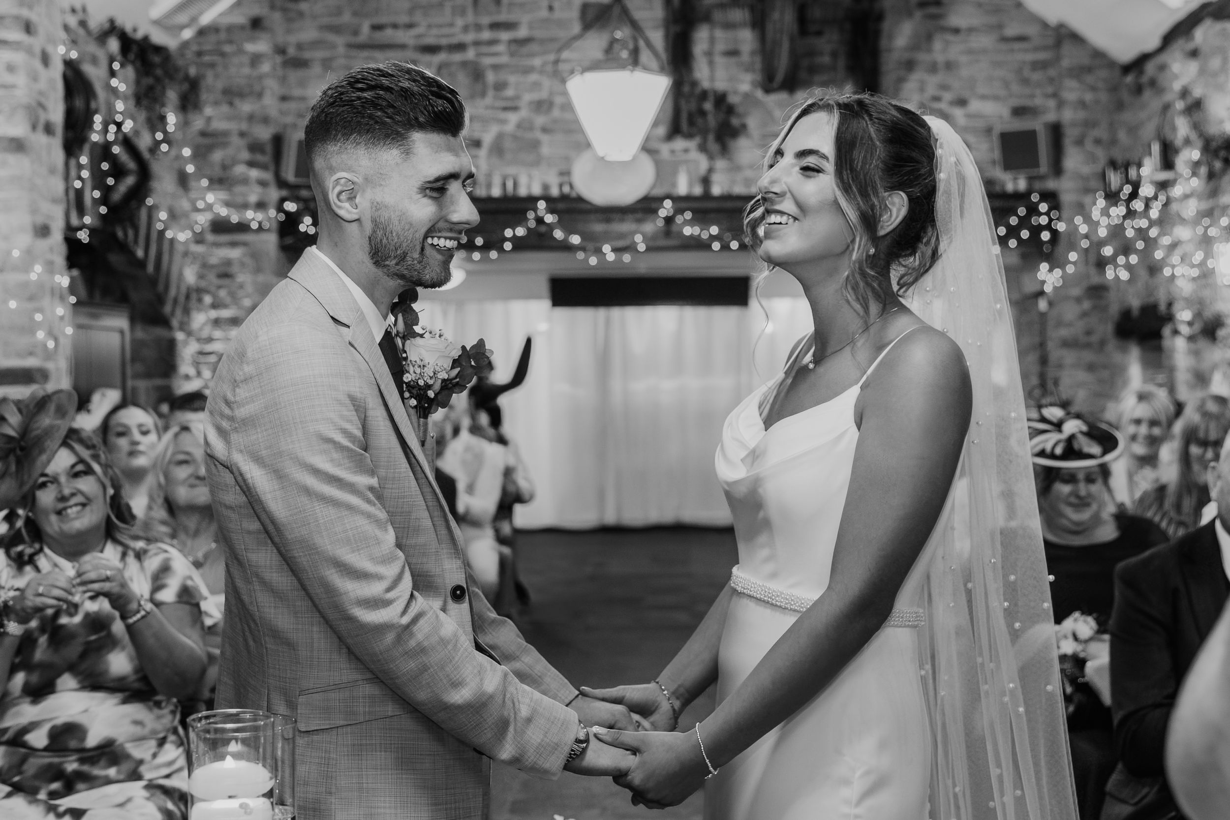 Black and white photo of a smiling bride and groom holding hands at their rustic wedding ceremony.