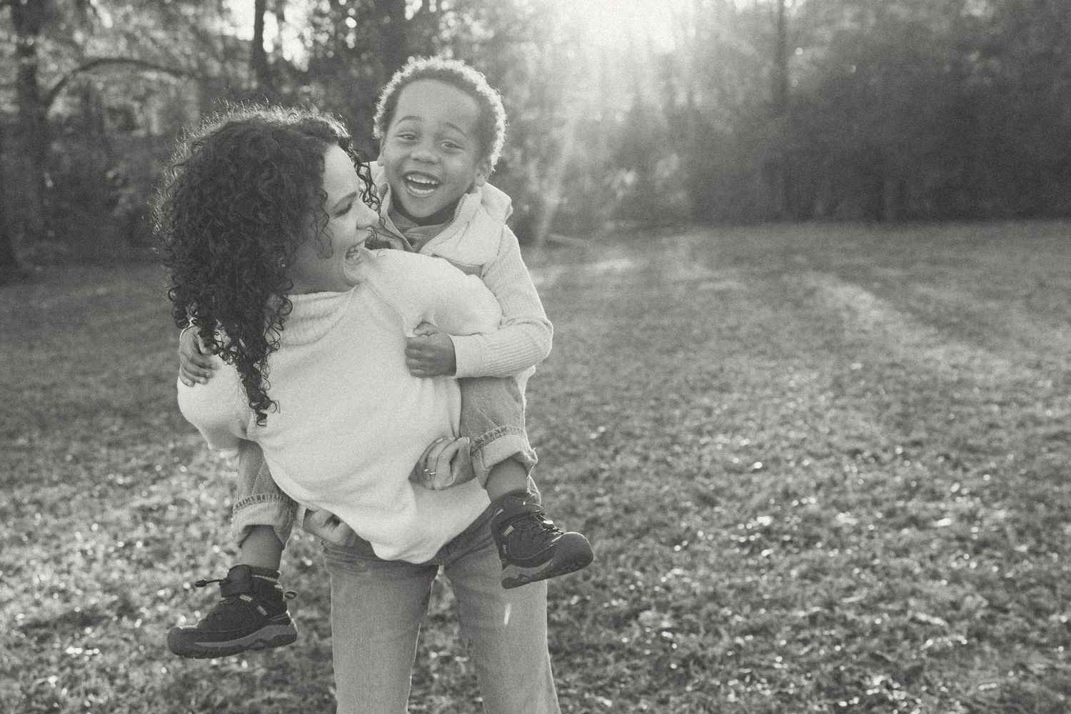 Black and white photo of a laughing child being held by a smiling woman outdoors in a sunlit park.