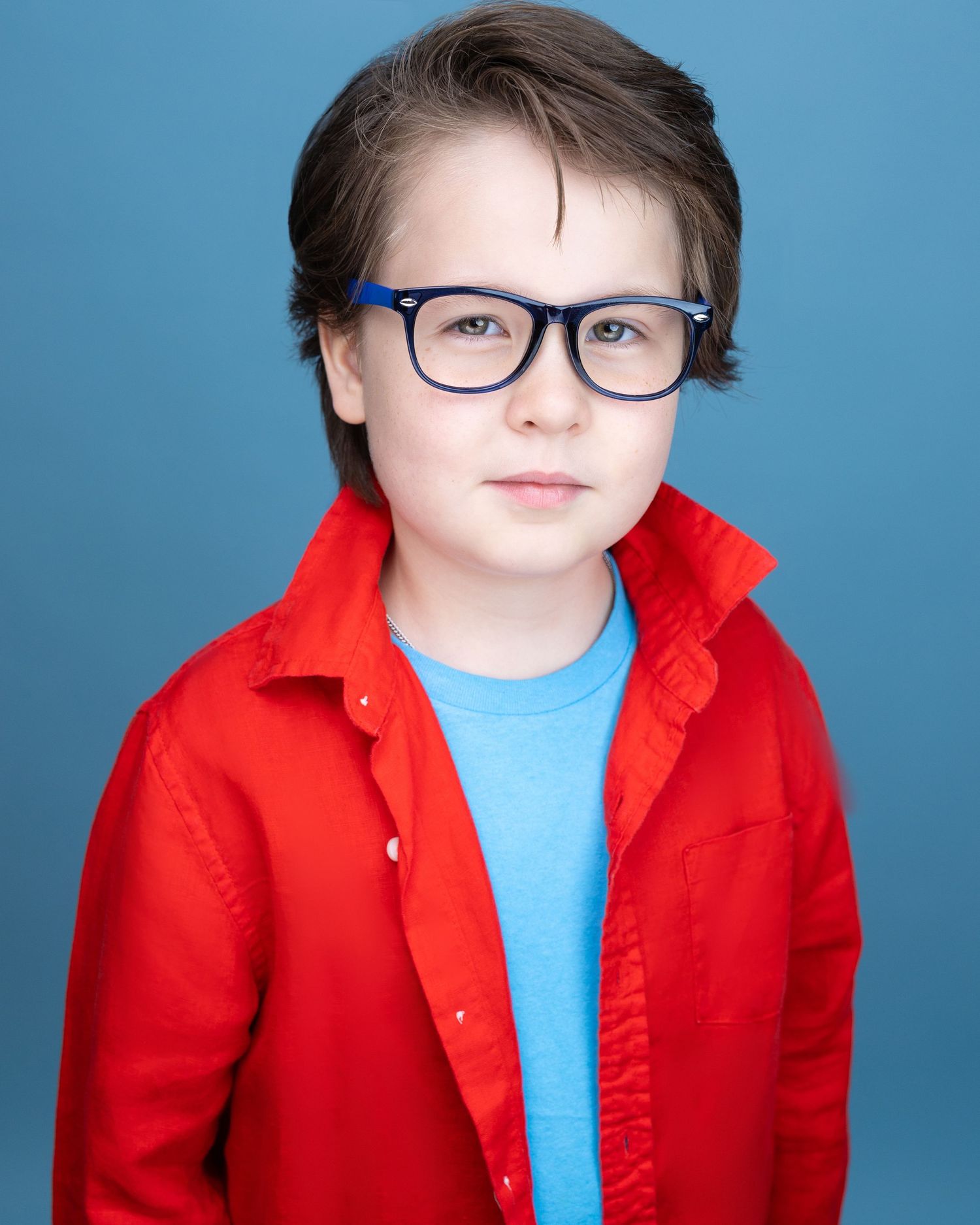 Young boy with blue glasses and red jacket poses for a professional headshot against a blue background.
