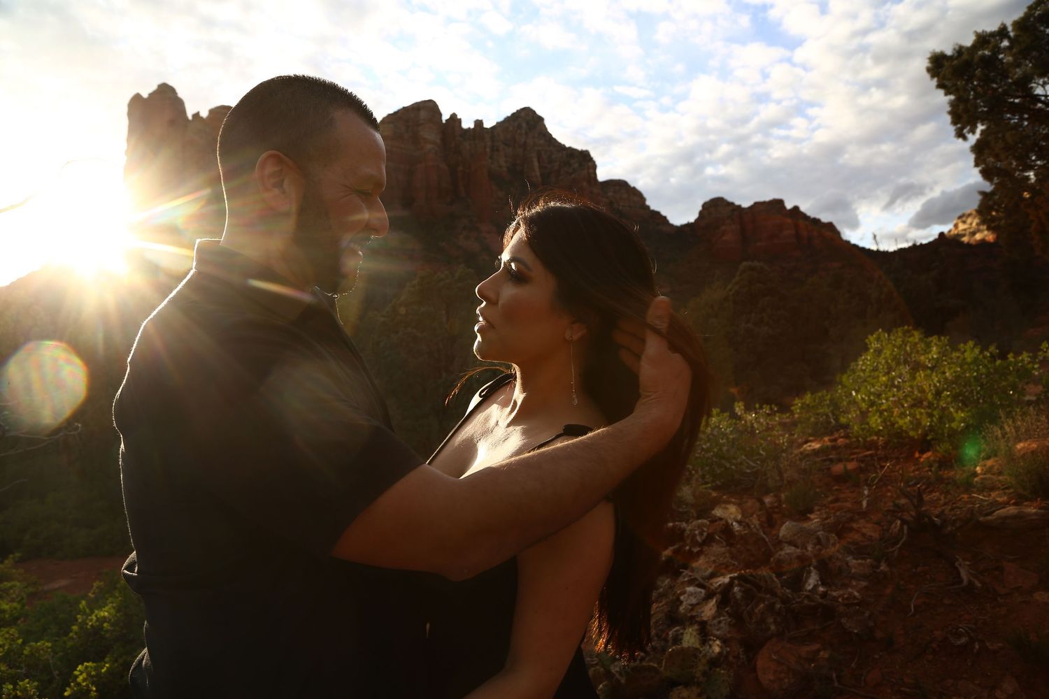 A couple embraces in golden sunset light against red rock formations in a scenic desert landscape.
