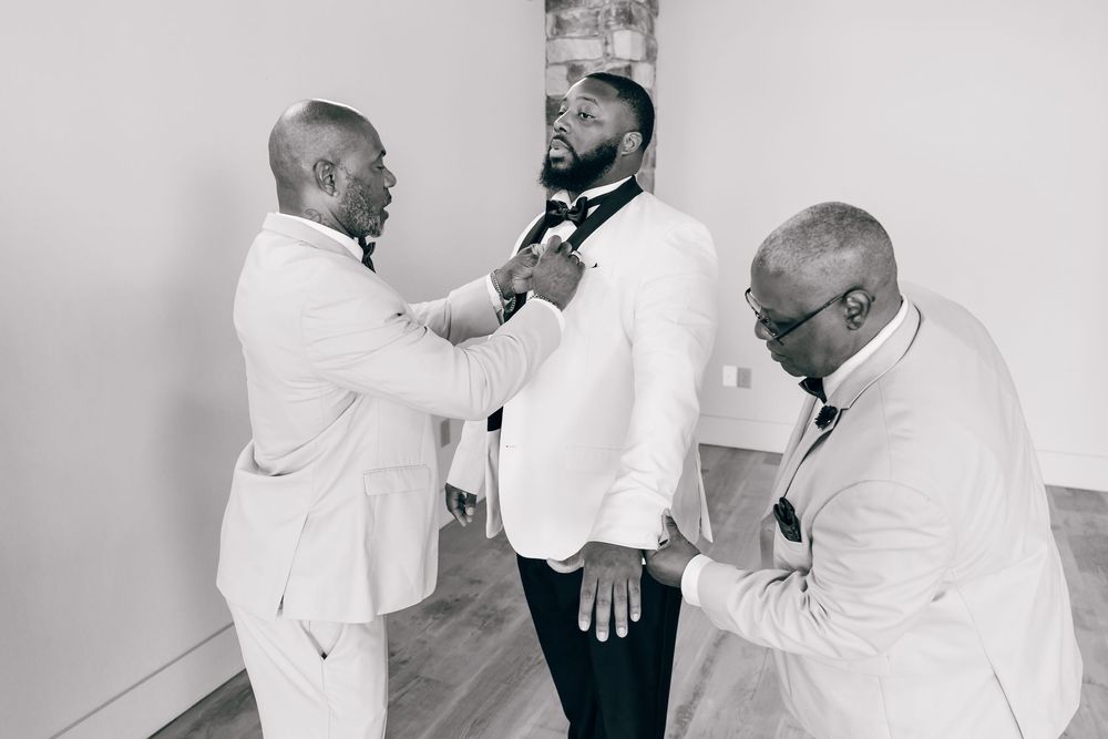 Black and white photo of groom being helped into his white tuxedo jacket by two groomsmen before wedding ceremony.