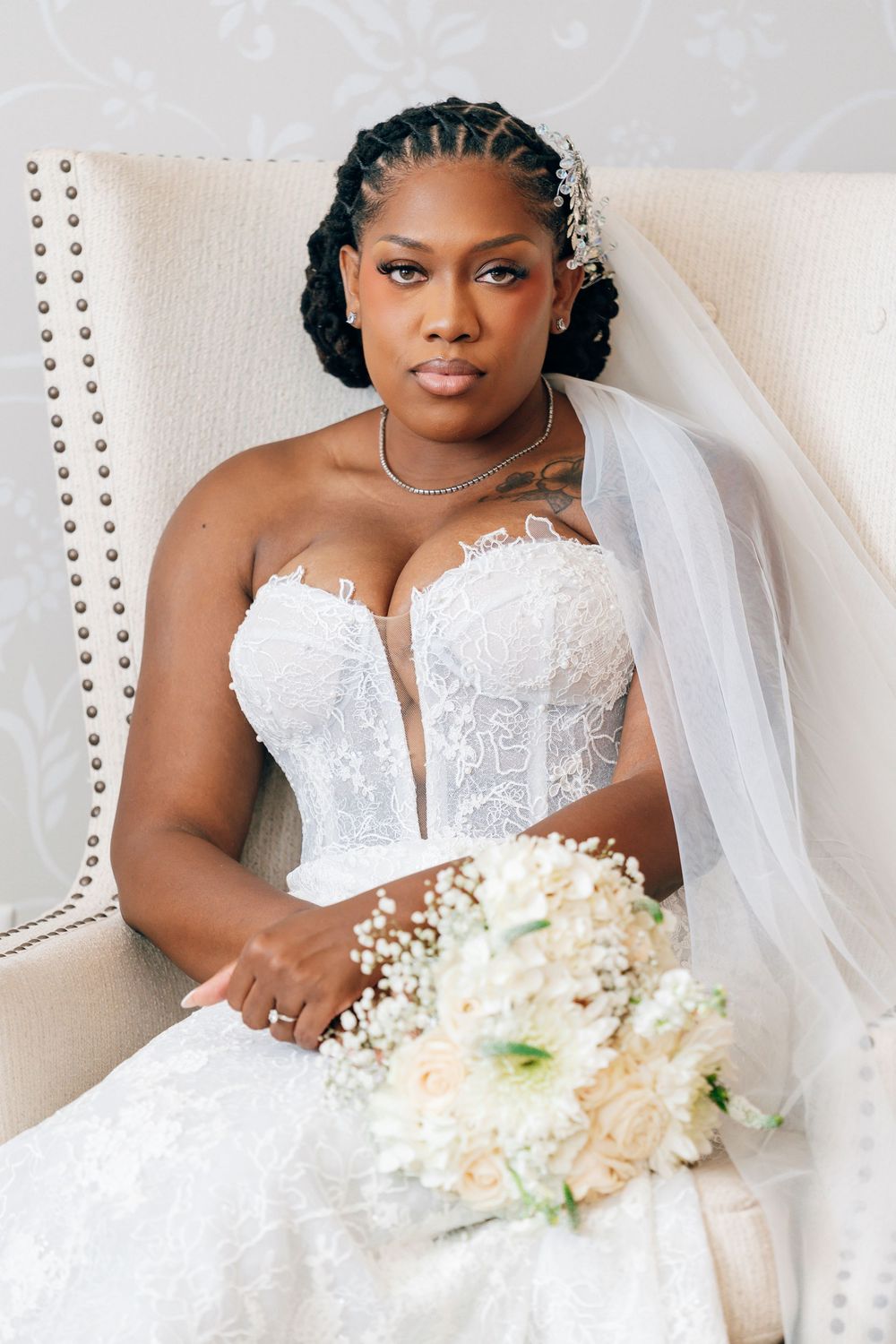 A beautiful Black bride in a strapless lace gown holds a white floral bouquet while seated in an elegant white chair.