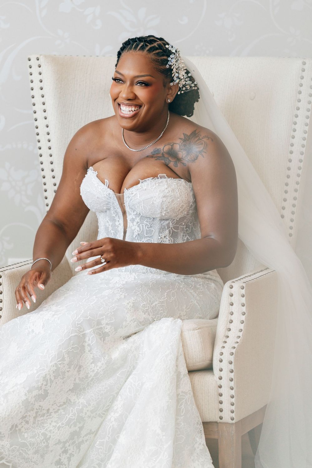 A radiant Black bride in a strapless lace mermaid gown sits smiling in a white tufted chair on her wedding day.
