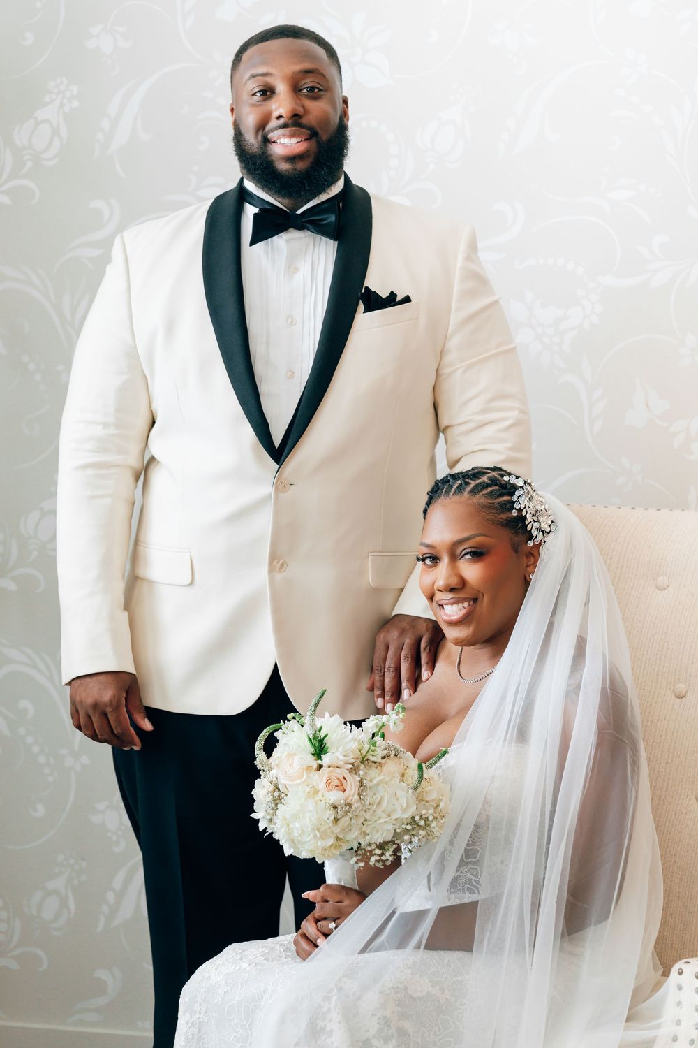 A Black groom in a white tuxedo stands beside his smiling bride in a lace gown holding a white floral bouquet.