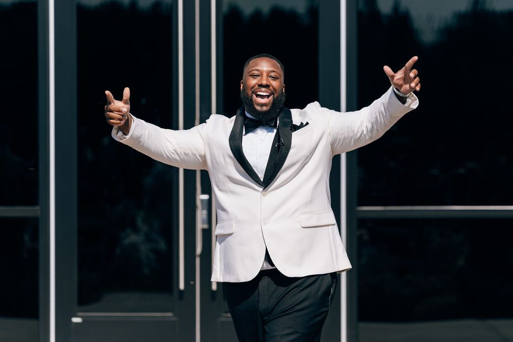 Joyful groom in white tuxedo jacket and bow tie celebrates with arms wide open outside modern building.