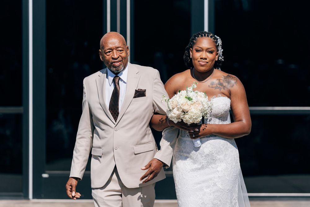 A Black bride in a strapless lace gown holding white flowers walks down the aisle escorted by an older man in a gray suit.