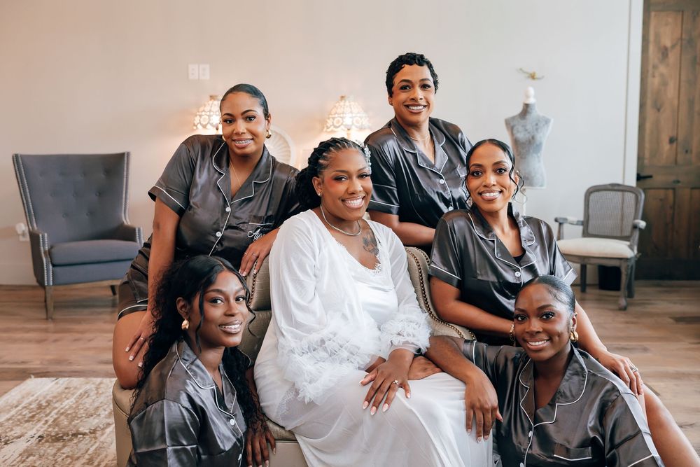 Bride in white robe poses with five bridesmaids in matching gray satin pajamas during wedding morning preparations.