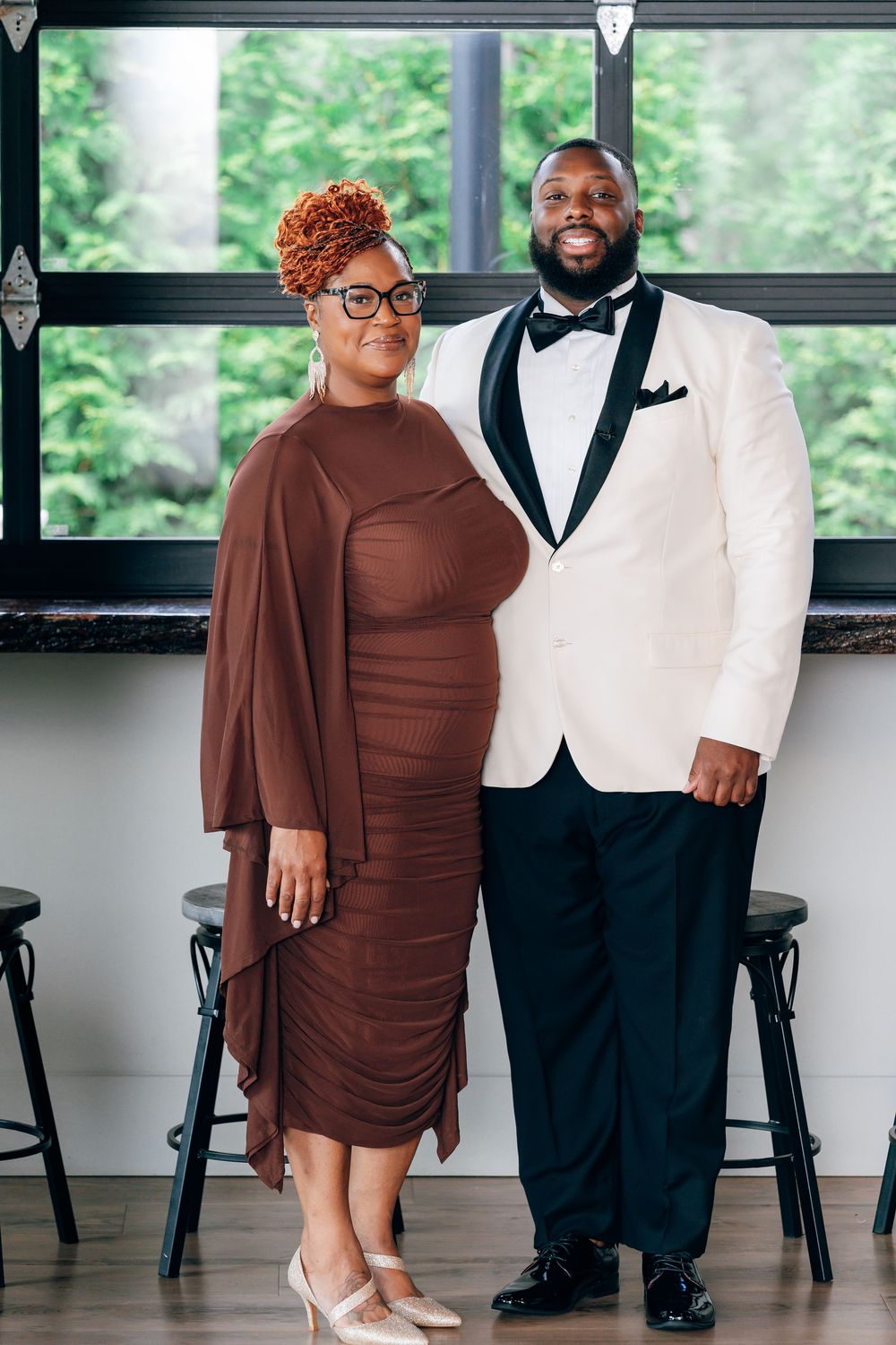 A well-dressed Black couple poses together, the woman in a brown midi dress and the man in a white tuxedo jacket with bow tie.