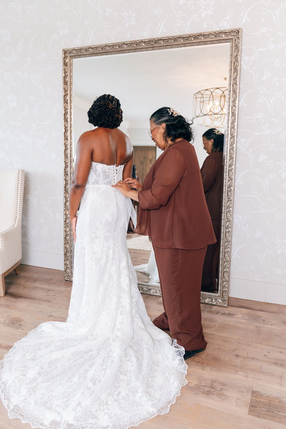 A bride in a white lace wedding gown is helped into her dress by a woman in a brown suit before a large mirror.