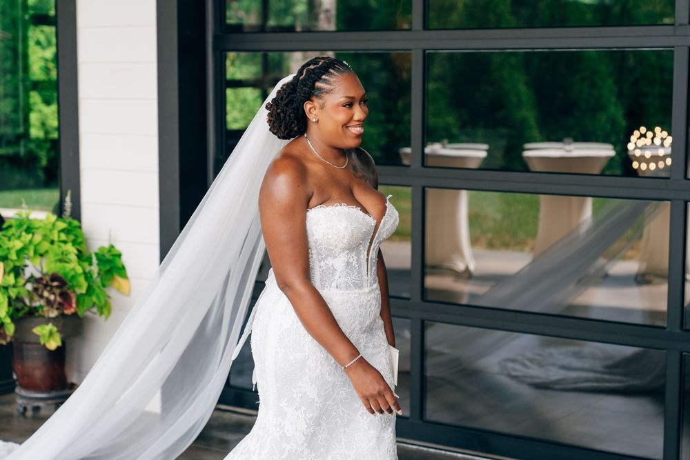 Bride in strapless lace wedding gown and flowing veil poses elegantly outdoors at a modern venue.
