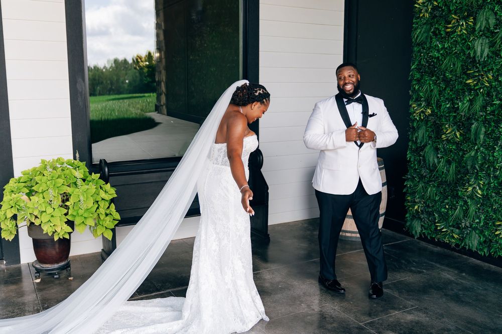 A bride in a white lace gown and veil shares a first look moment with her groom in a white tuxedo jacket.