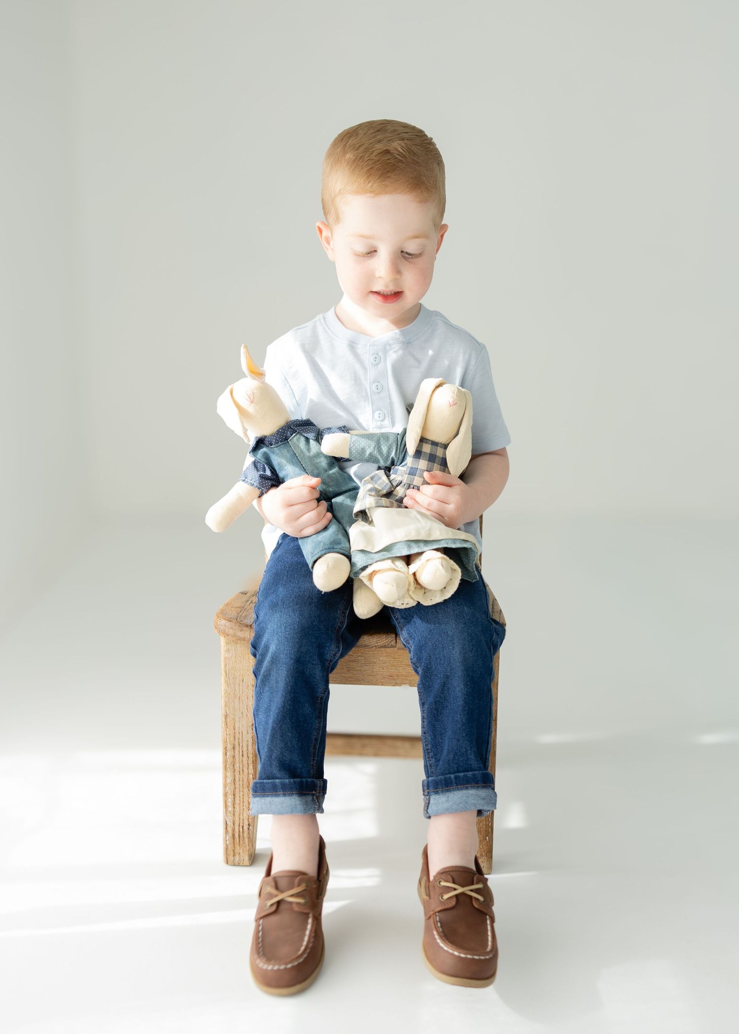 Young redhead boy sitting on wooden stool holding stuffed animal toys, wearing jeans and brown loafers.