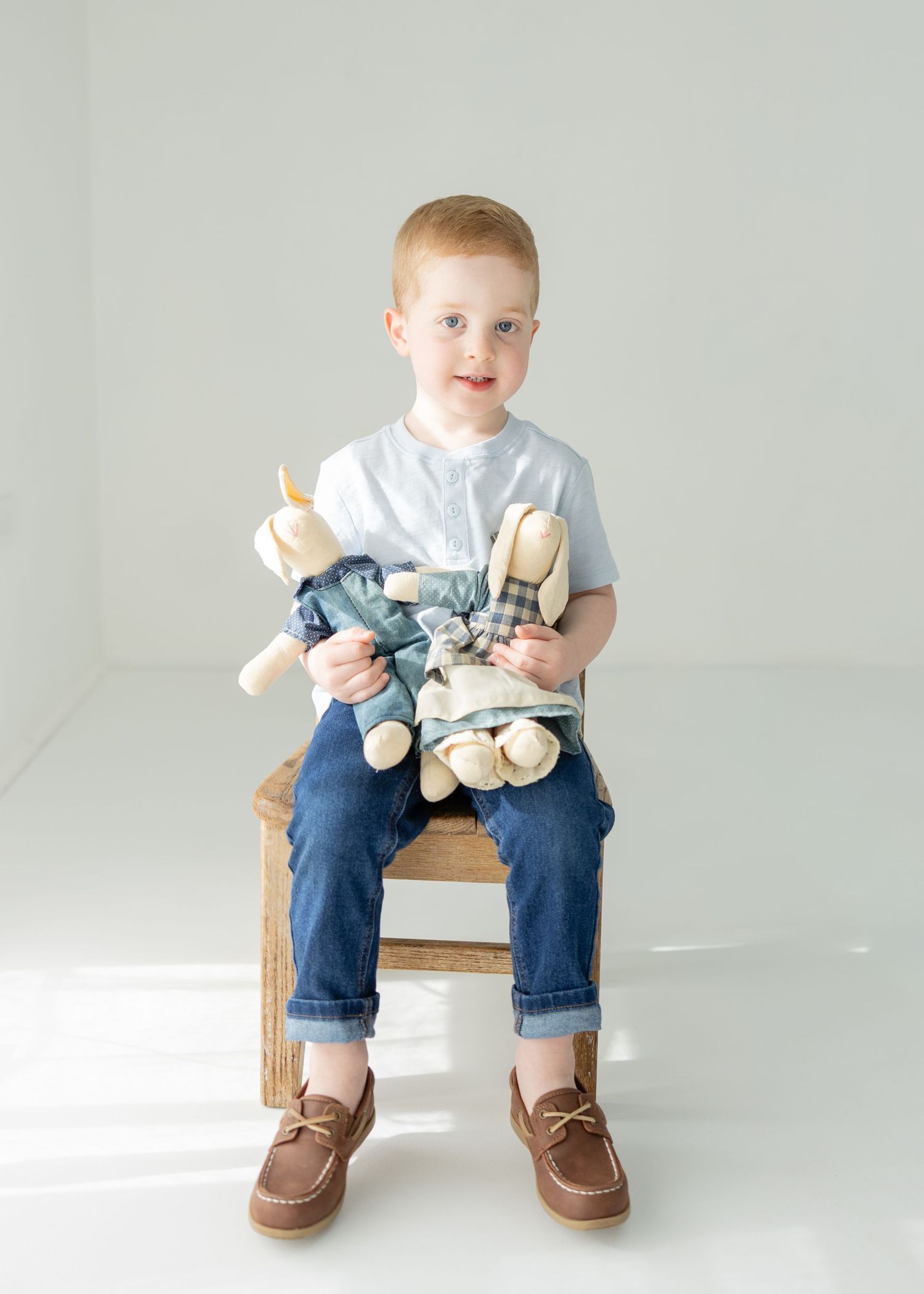Red-haired boy sitting on wooden stool holding stuffed animal toys, wearing blue jeans and white shirt.