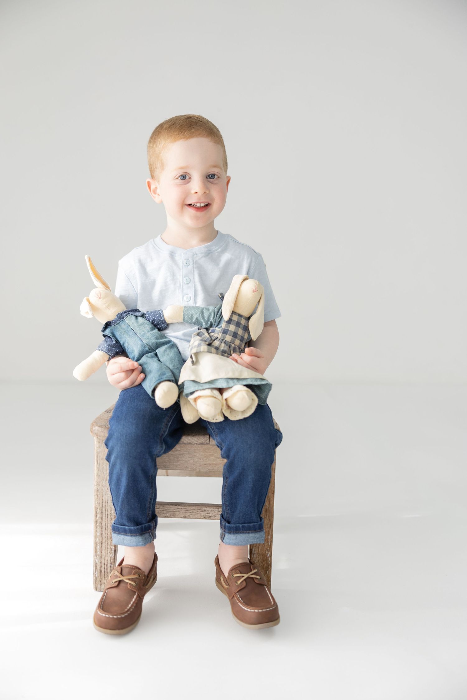 Smiling redhead toddler boy sitting on wooden stool holding stuffed bunny toys, wearing blue shirt and jeans.