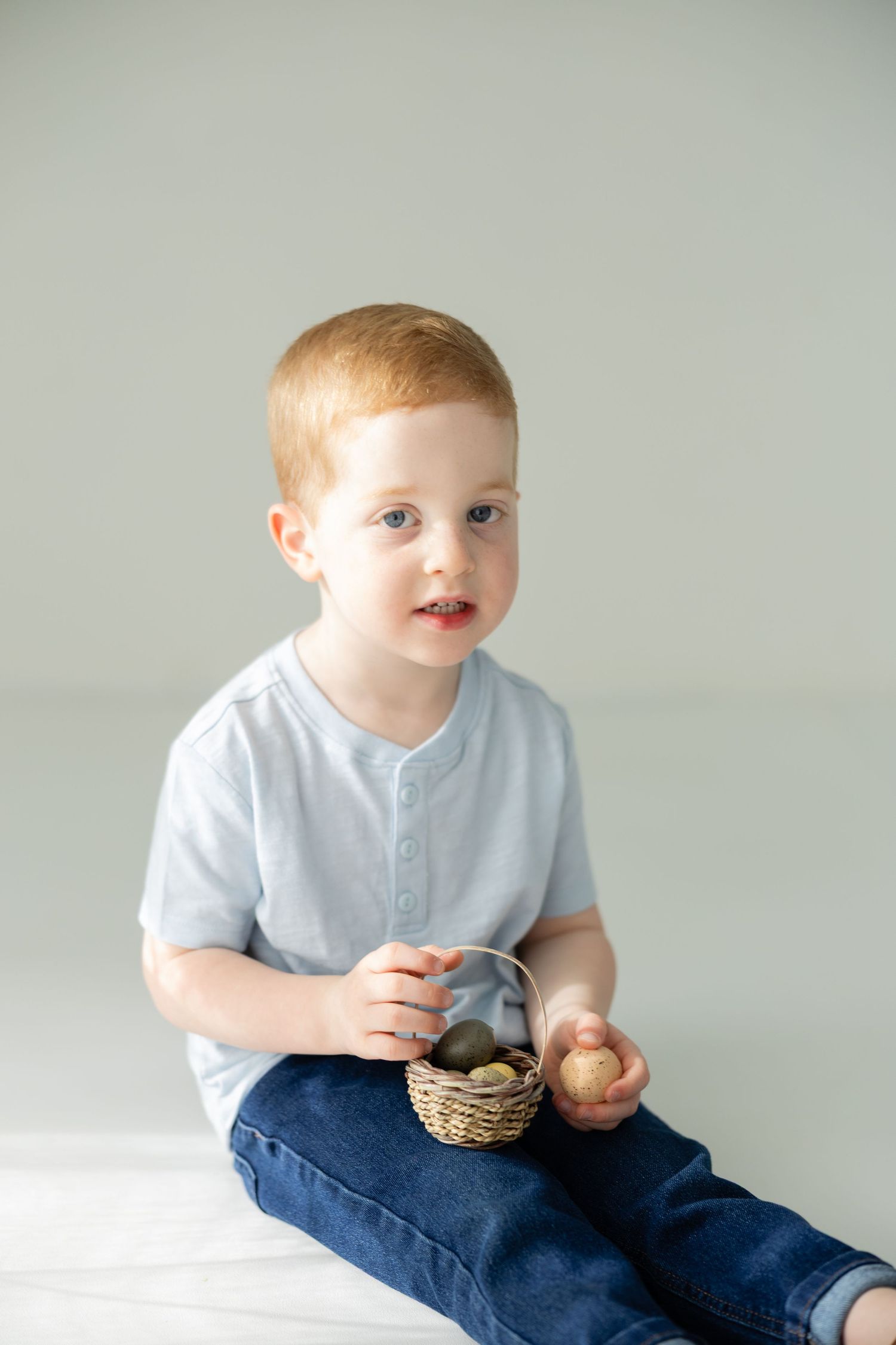 Cute redhead toddler boy sitting and holding a small wicker basket with Easter eggs on a white background.