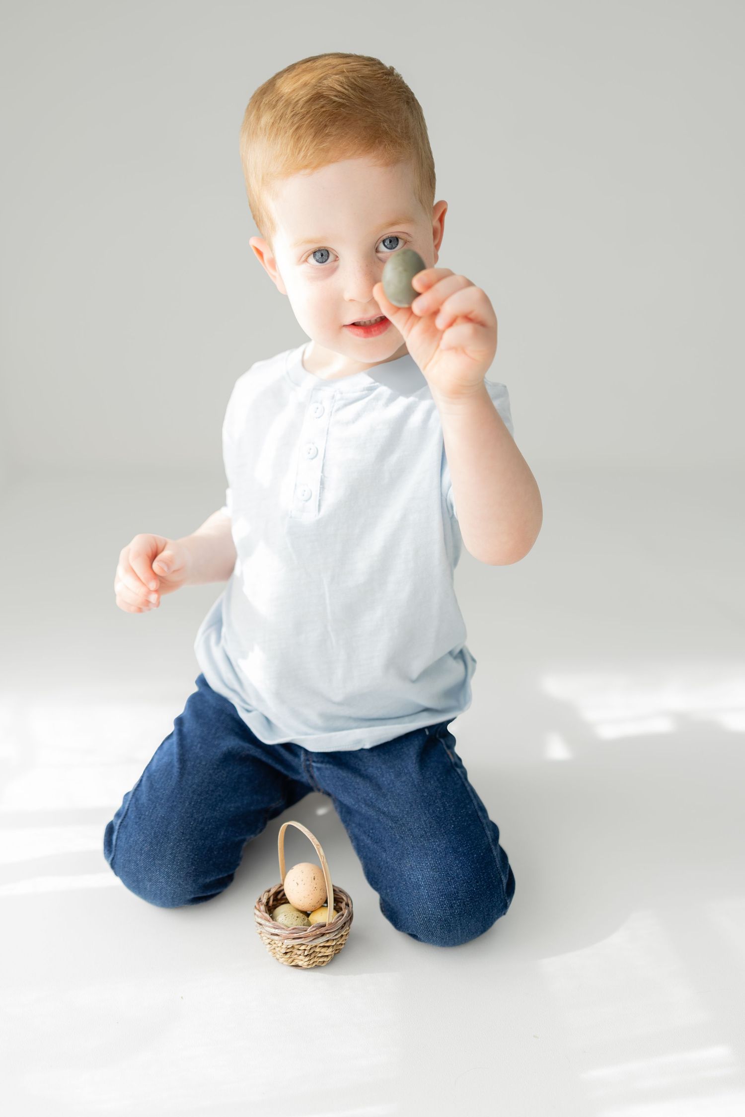 Adorable toddler boy holding an egg near a small wicker basket on white background.