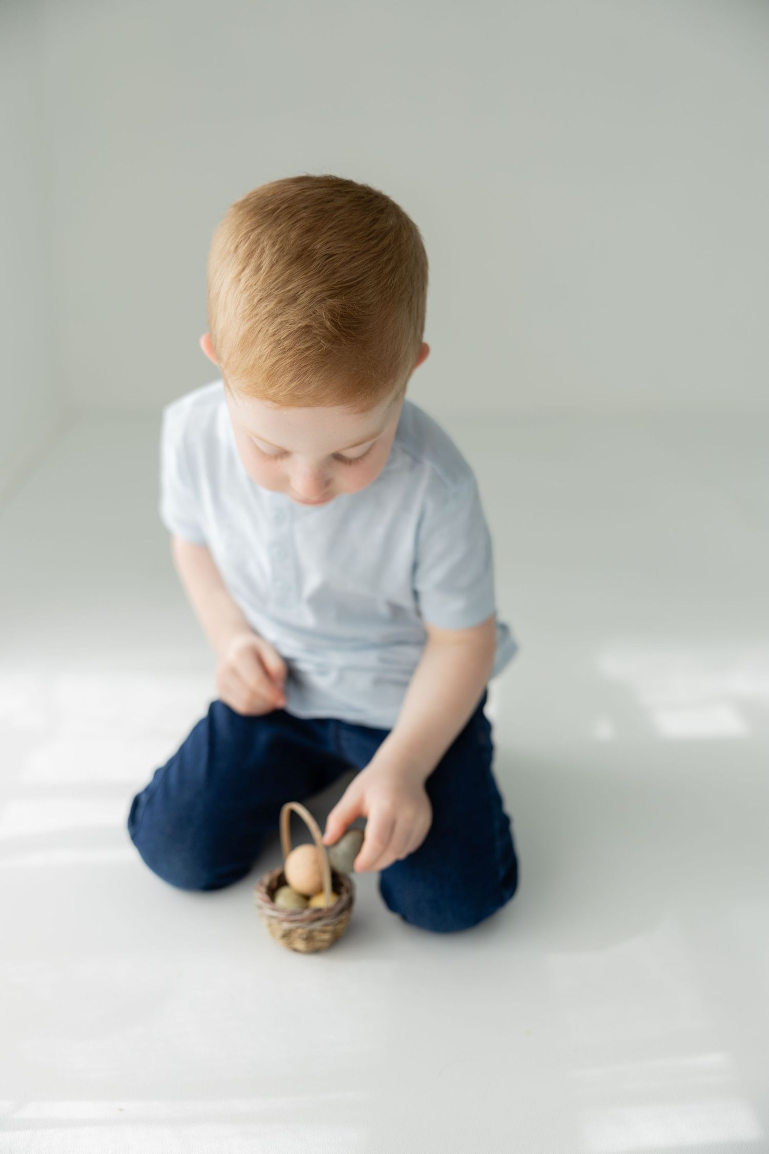 Red-haired toddler boy sitting on white floor holding a small wicker basket with Easter eggs.