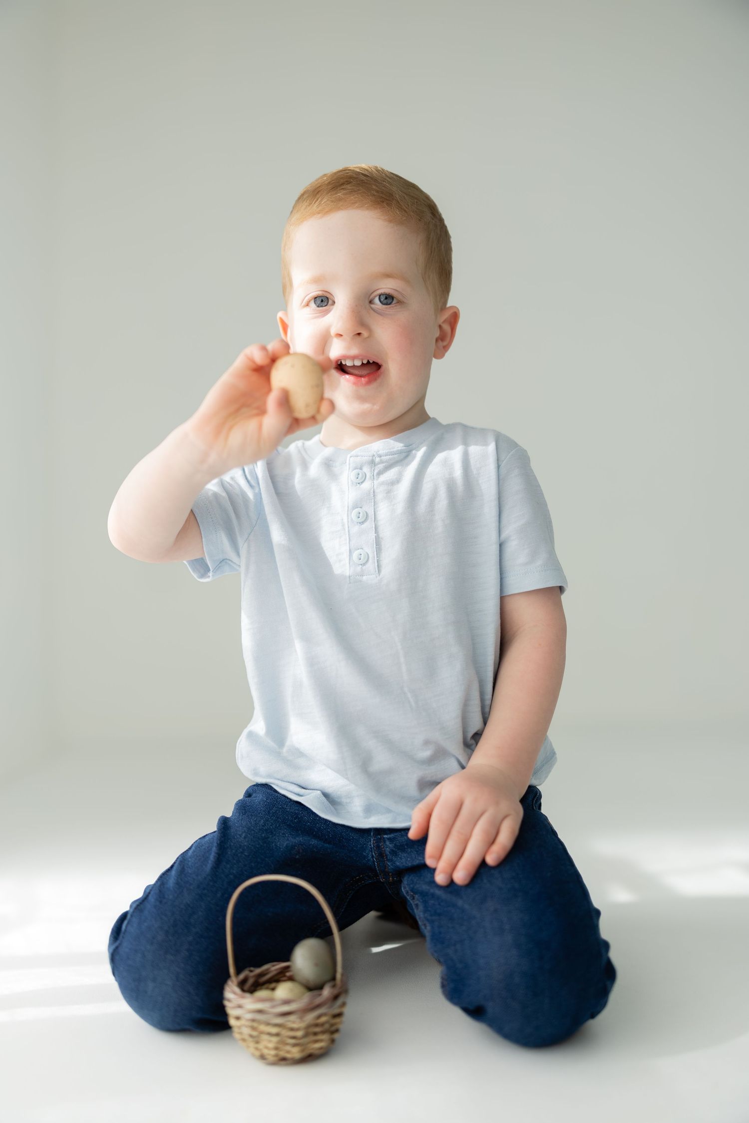 Happy redhead toddler boy holding an Easter egg beside a small wicker basket with eggs on white background.