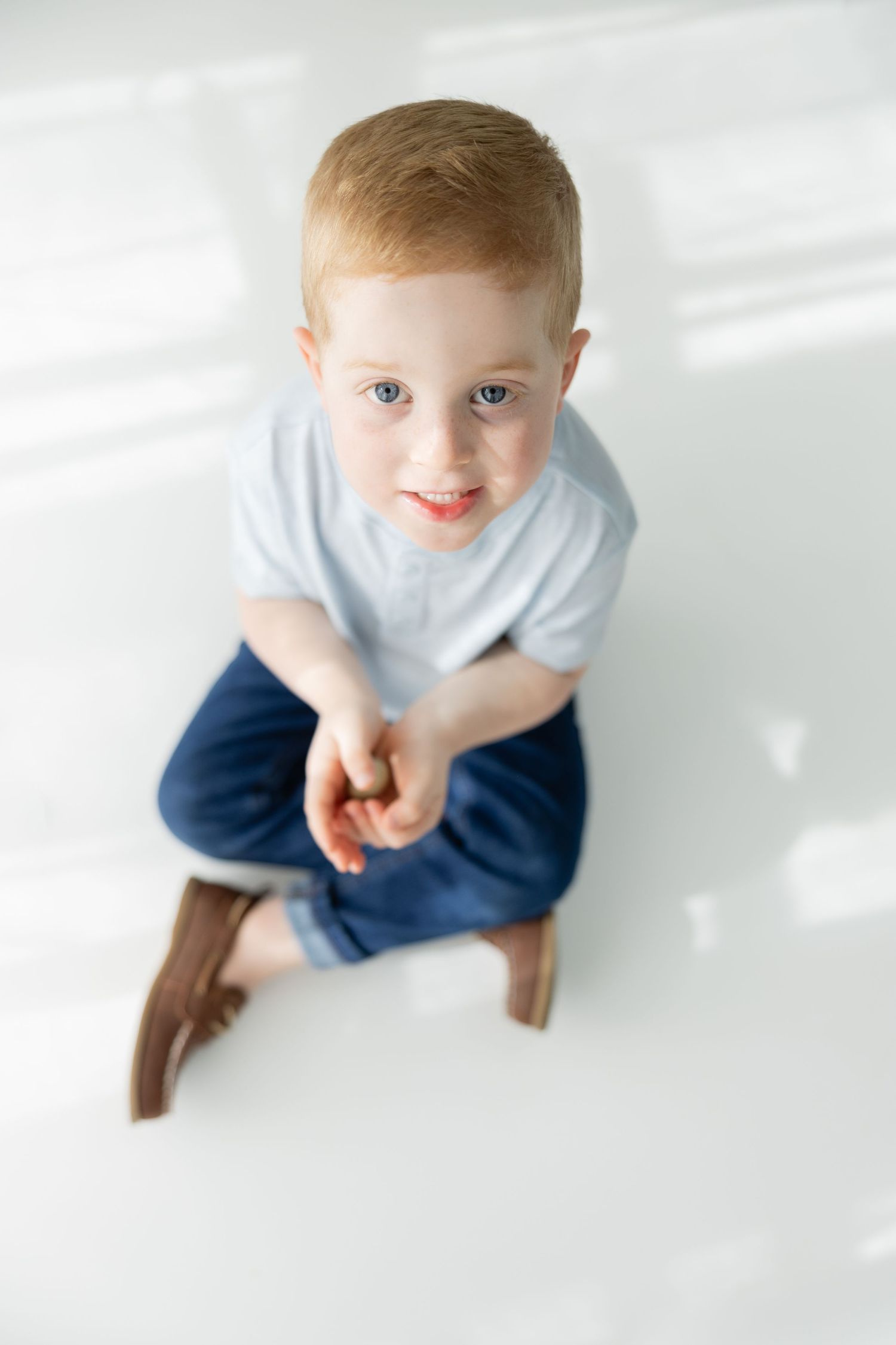 Smiling redhead toddler boy sitting cross-legged on white floor, wearing light blue shirt and navy shorts.