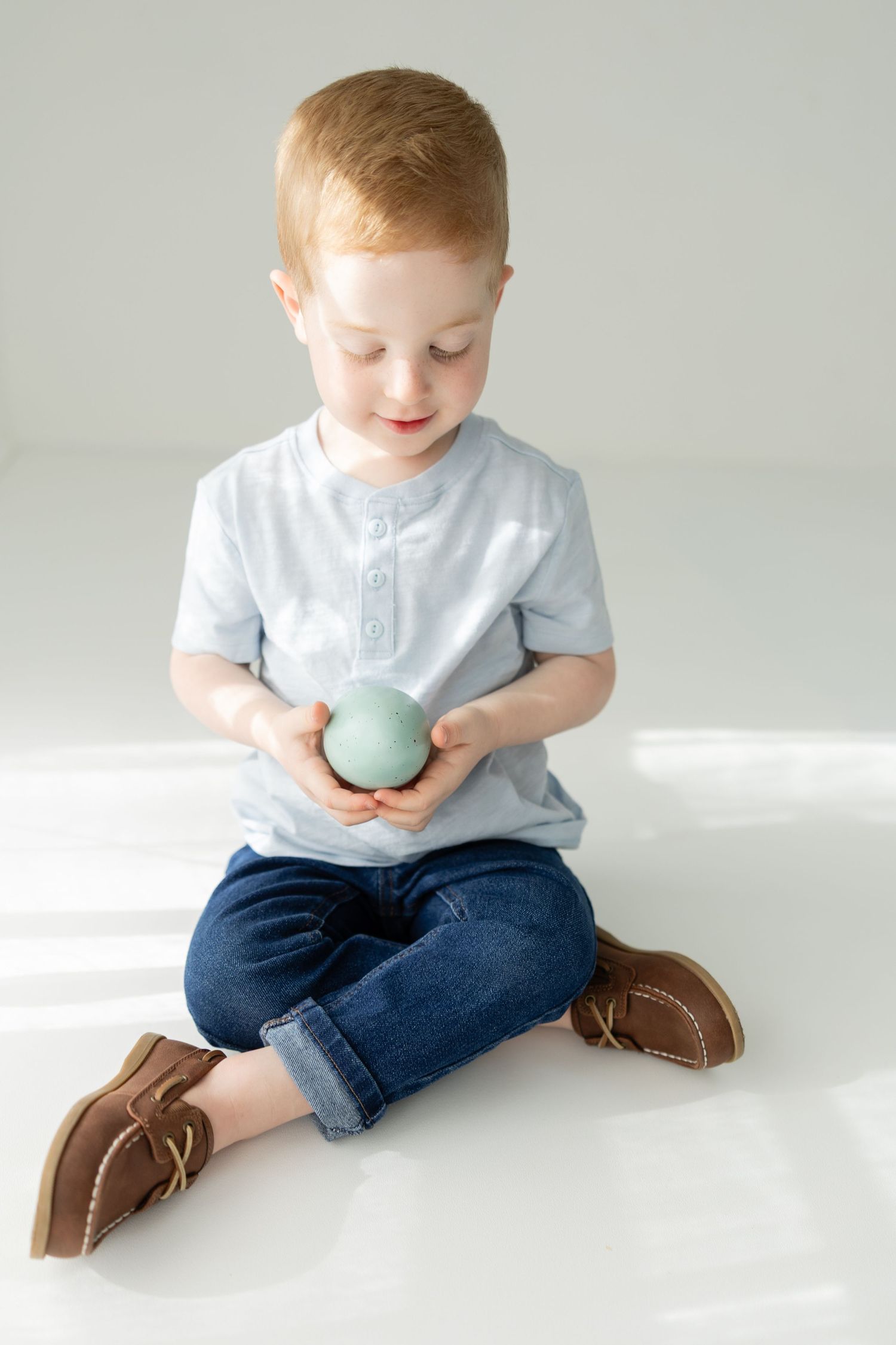 Adorable redhead toddler boy sitting cross-legged holding a mint green ball, wearing blue jeans and brown shoes.