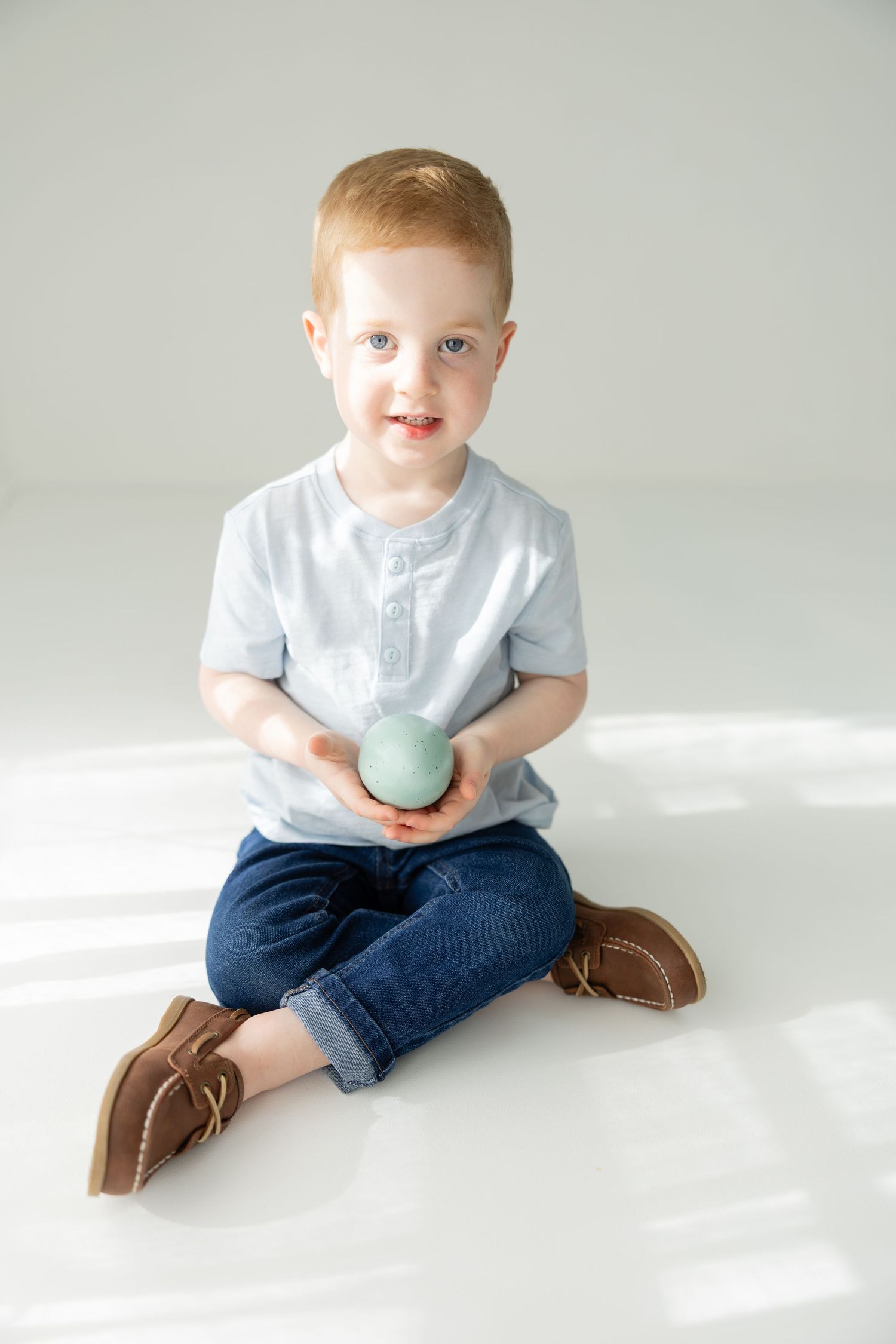 Smiling toddler boy with red hair sitting on white floor holding a green ball, wearing blue jeans and a gray shirt.