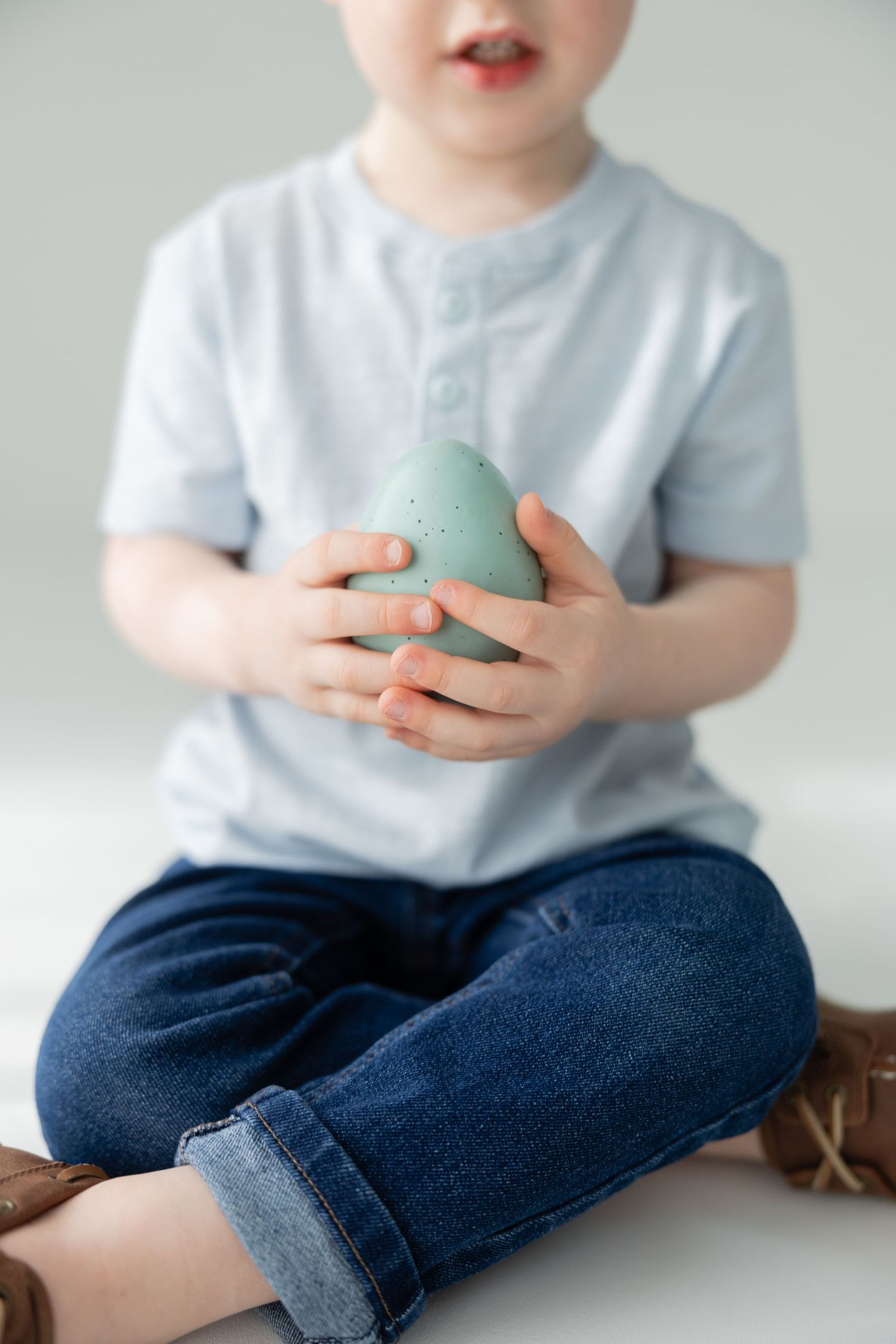 Young toddler boy sitting cross-legged holding a mint green speckled Easter egg with both hands.